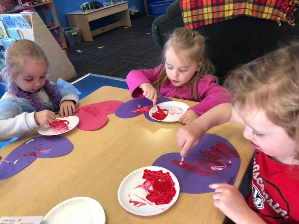 Three children painting heart shapes in a classroom