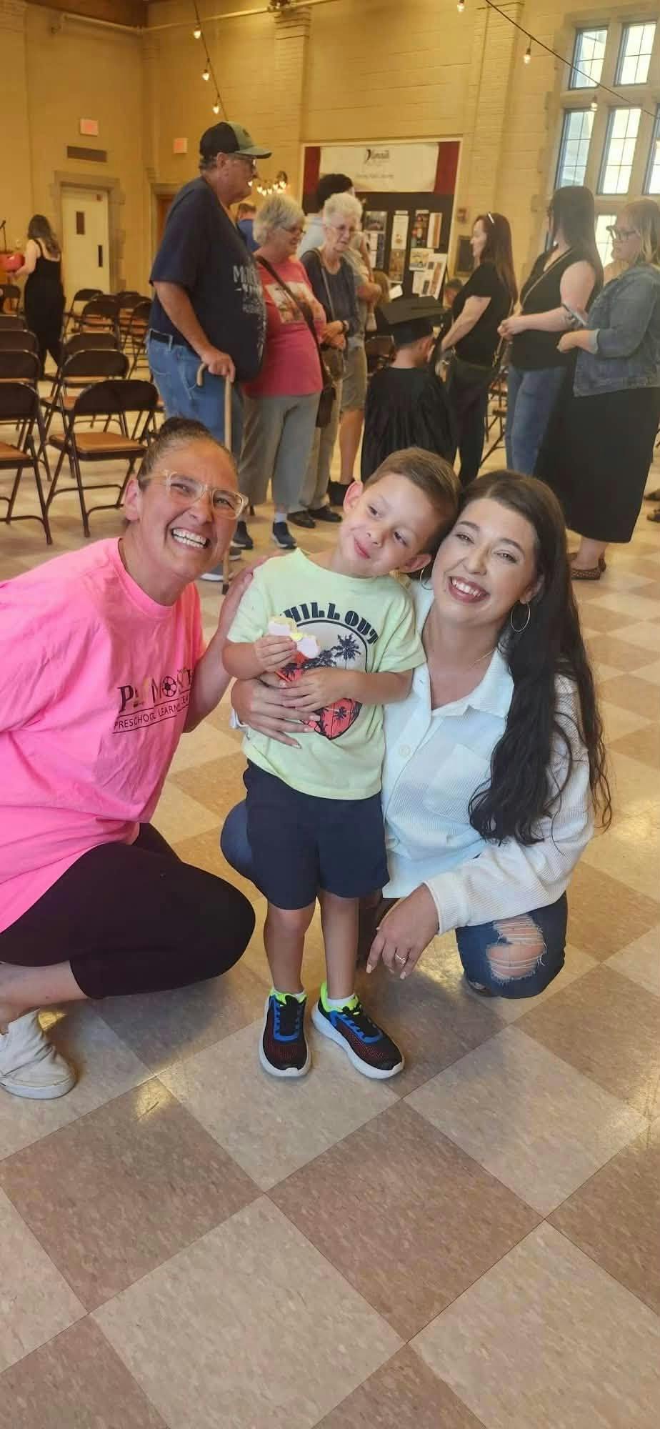 Two people crouching beside a child in a room with other people in the background