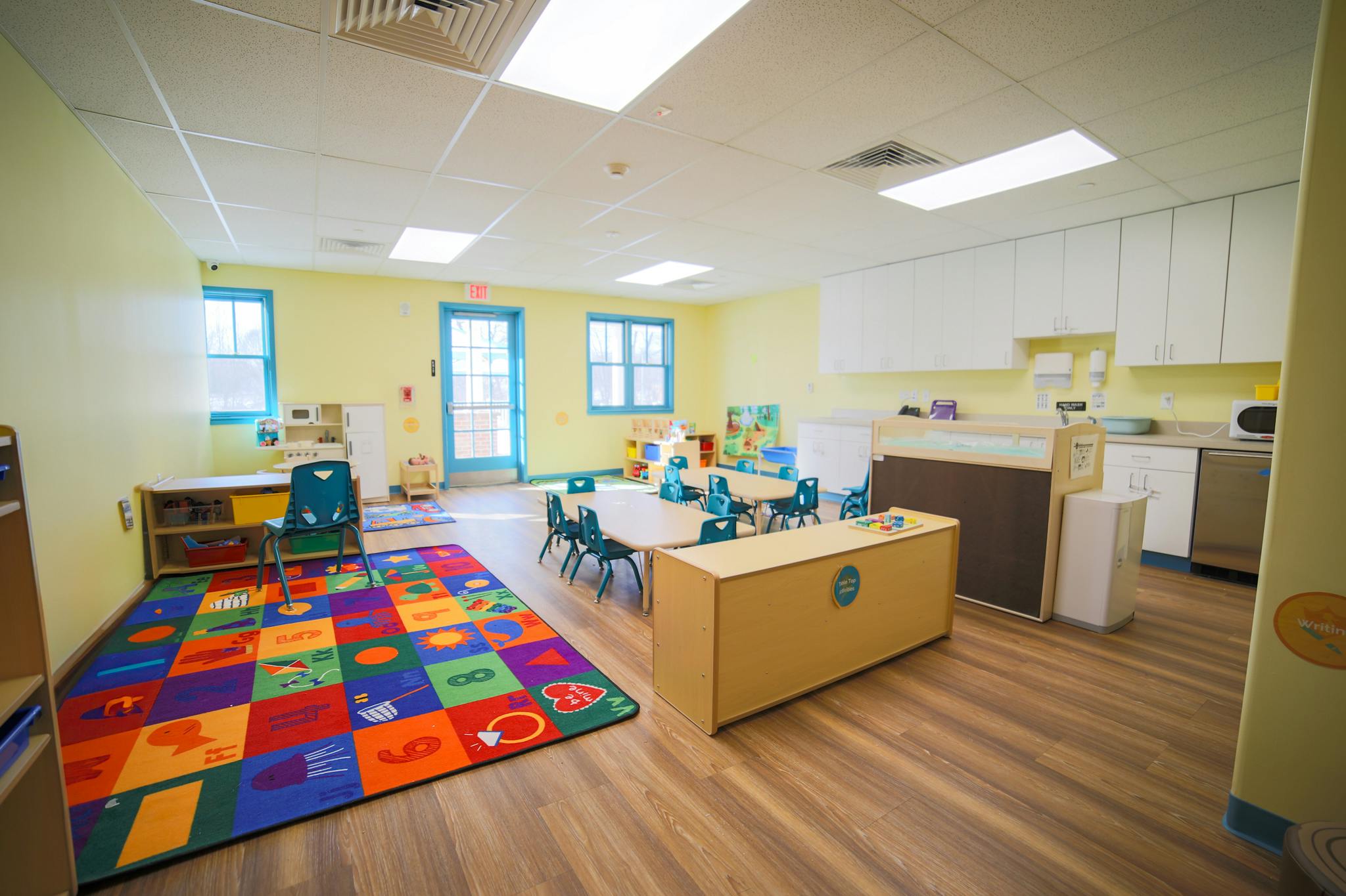 Empty classroom with colorful rug, small tables and chairs, and kitchen area