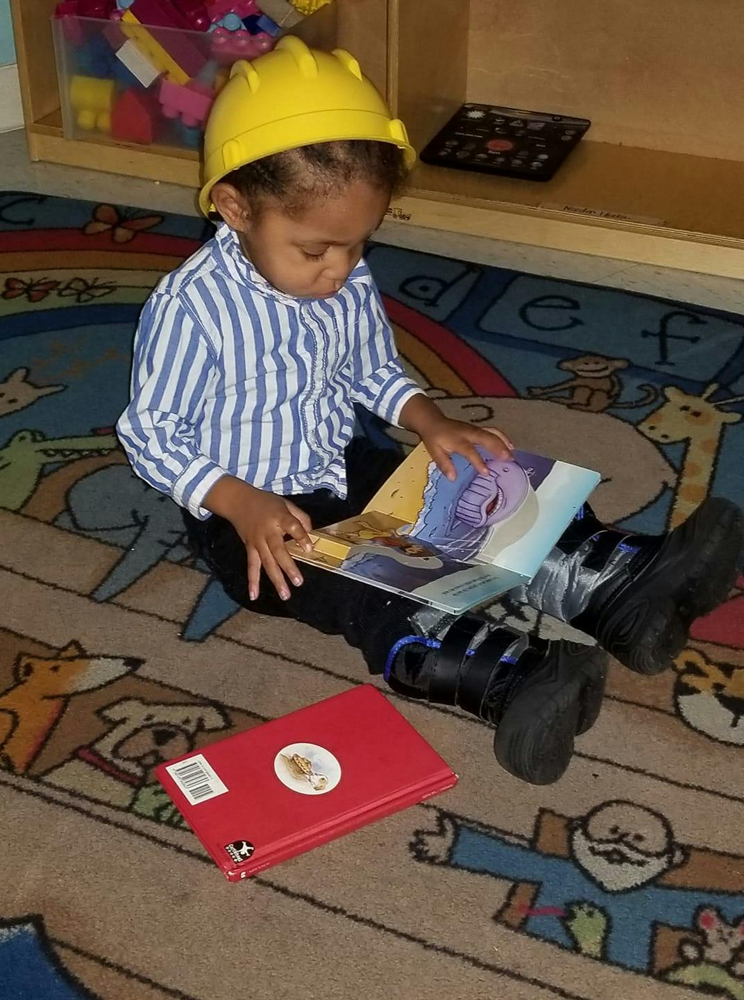 Child in a yellow hard hat sitting on a rug and looking at a book