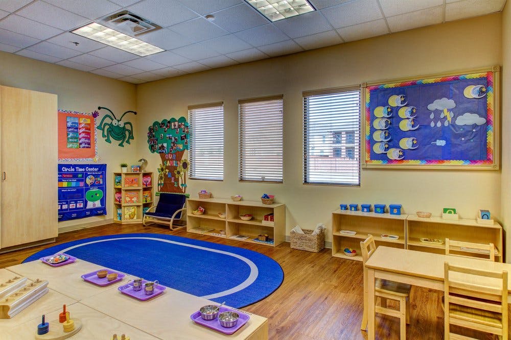 Classroom with a blue oval rug, tables, chairs, and shelves with learning materials
