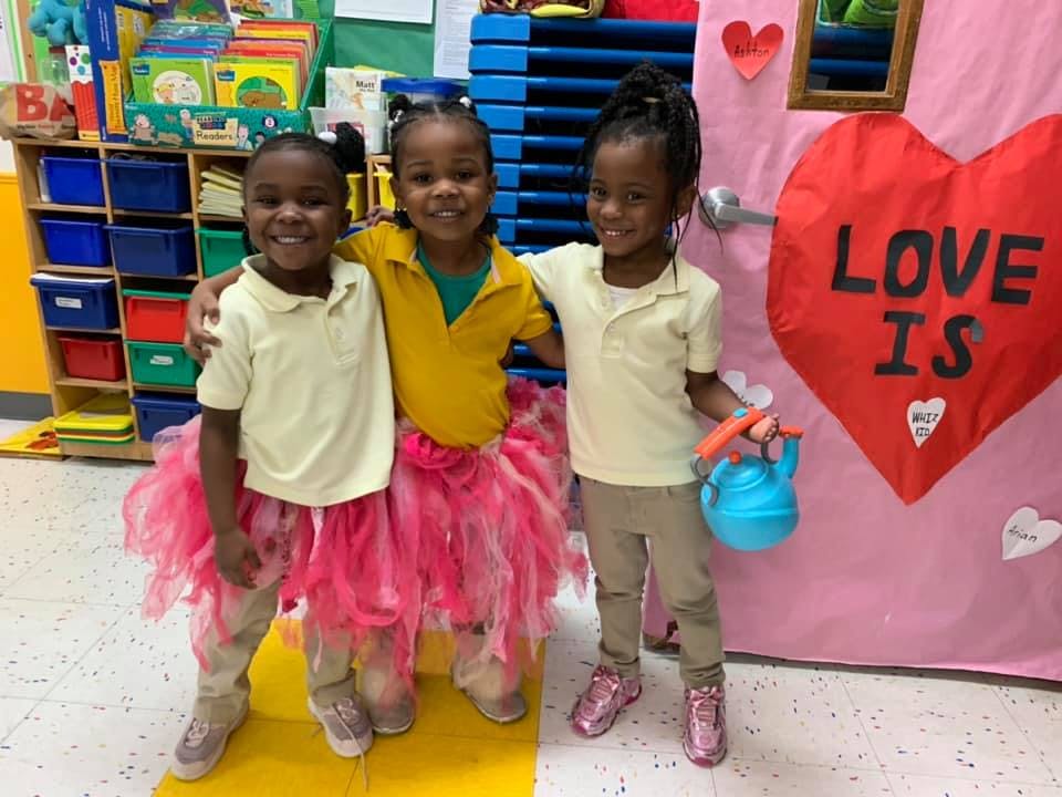 Three children standing together, two wearing pink tutus