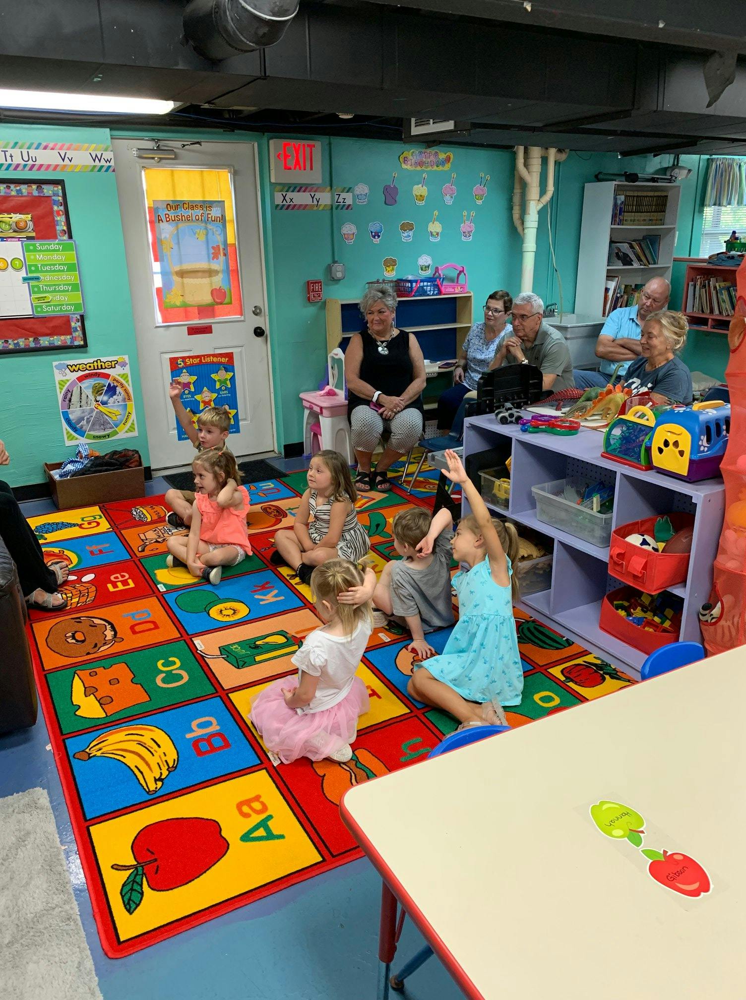 Children sitting on a colorful alphabet rug in a classroom with adults watching