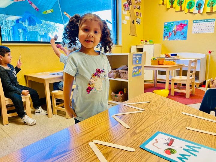 Child standing in a classroom with other children and tables