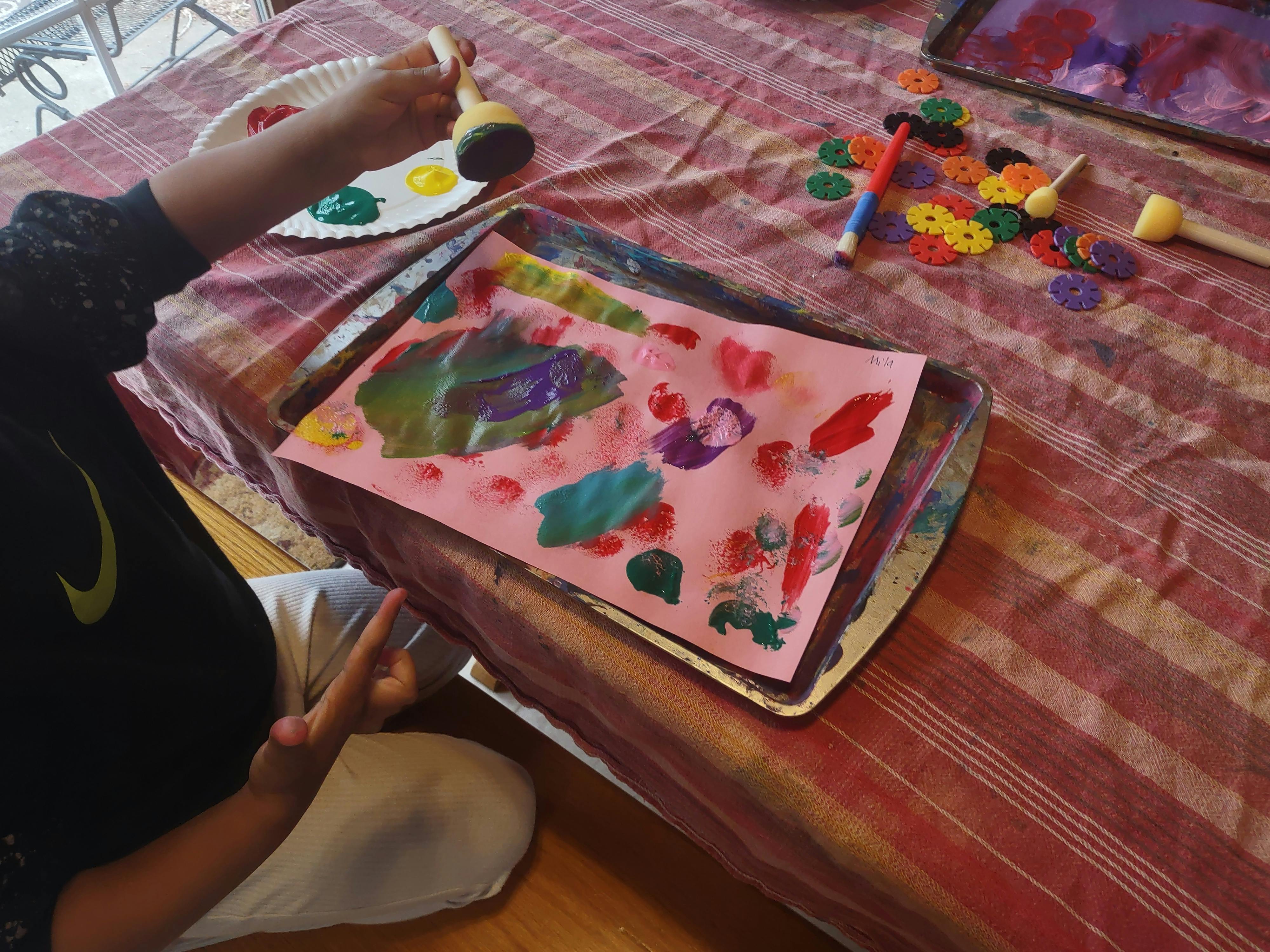 Child's hand holding a round sponge paintbrush over a colorful painting