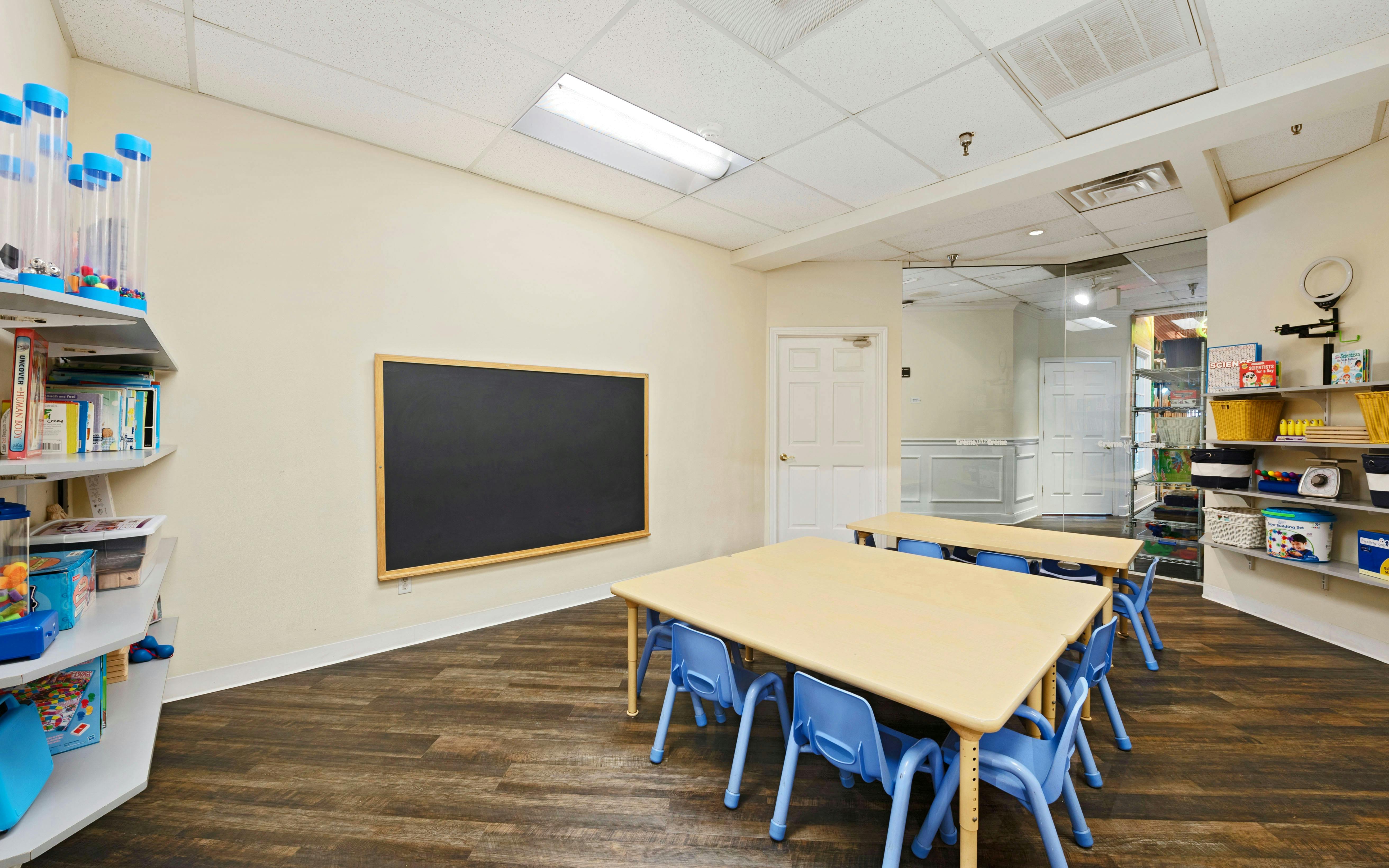 Empty classroom with two small tables and chairs, chalkboard, and shelves of toys