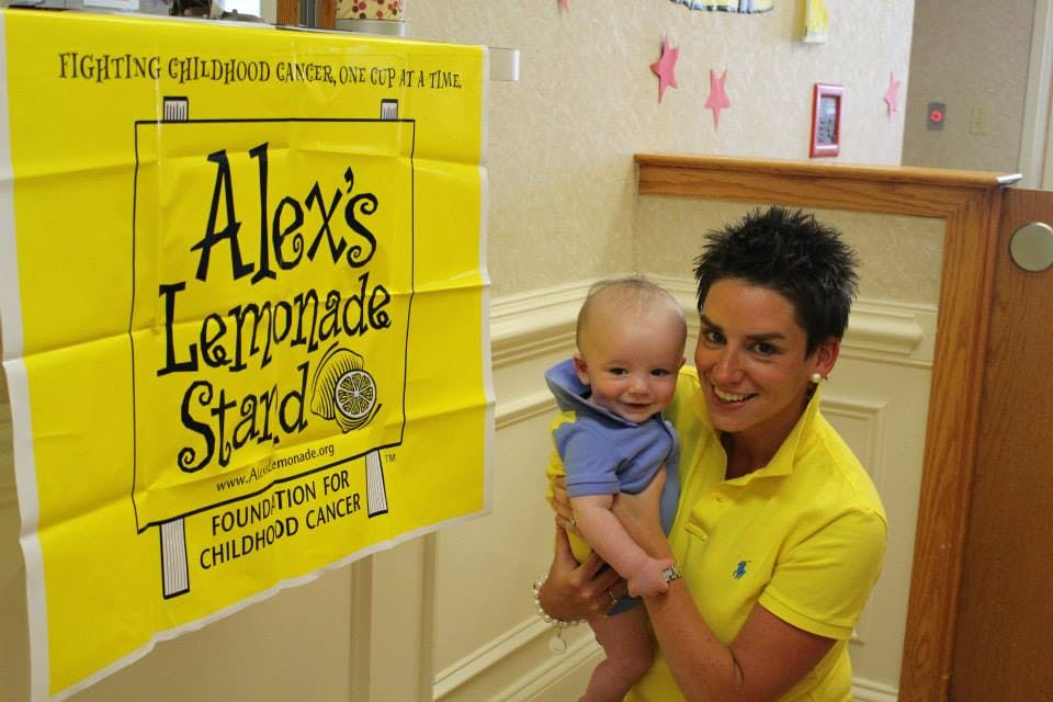 Person holding a baby next to an Alex's Lemonade Stand sign