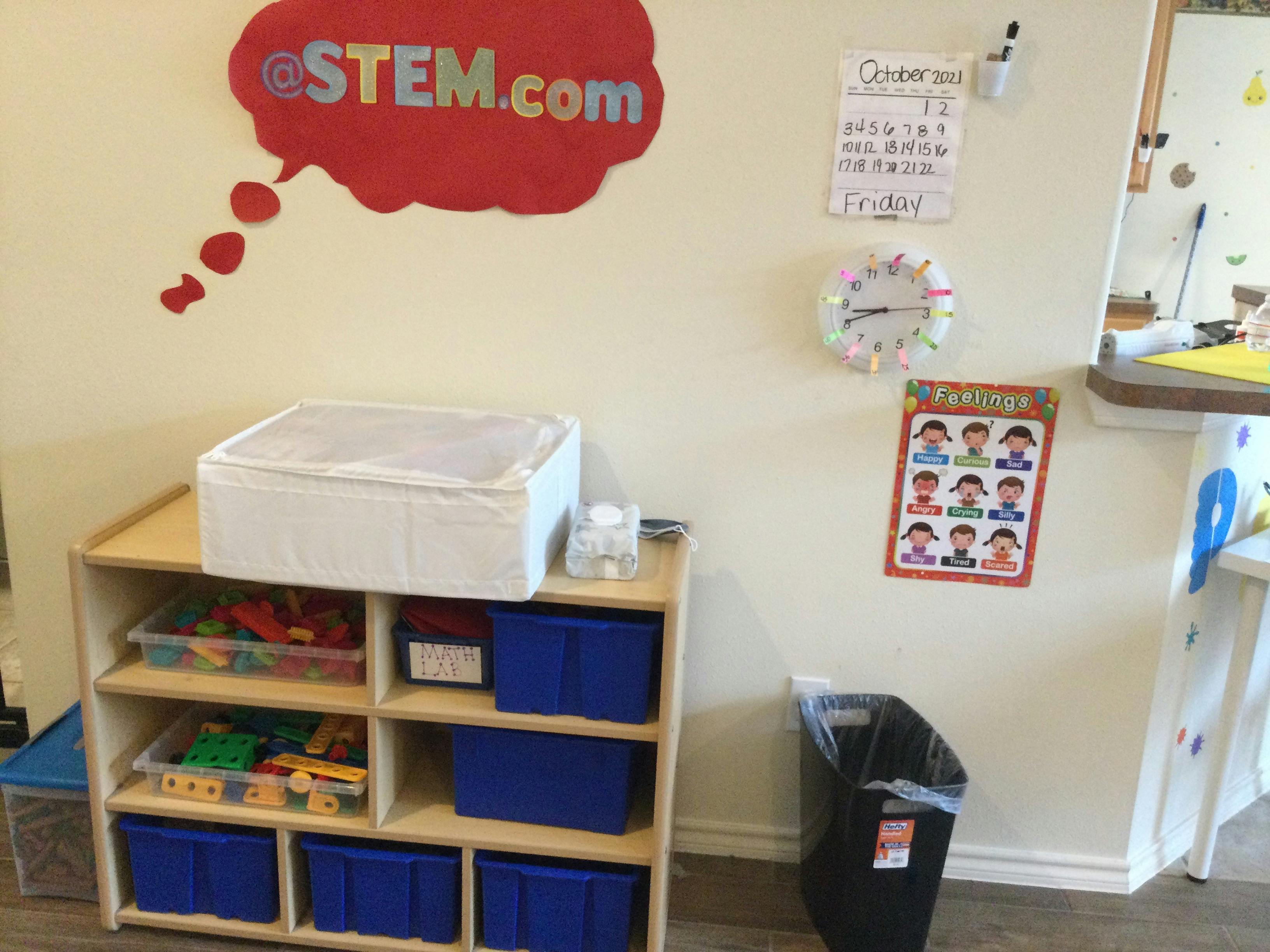 Classroom wall with a STEM.com sign, calendar, clock, and a shelf with toys