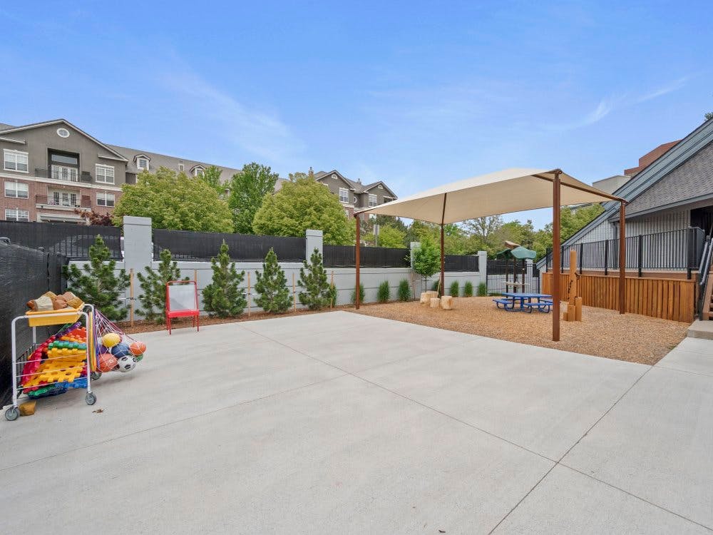 Outdoor play area with a sunshade over wood chips, a picnic table, and a cart of toys