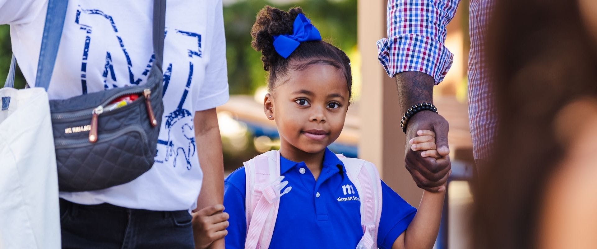 Child wearing a backpack and blue uniform, holding hands with two people