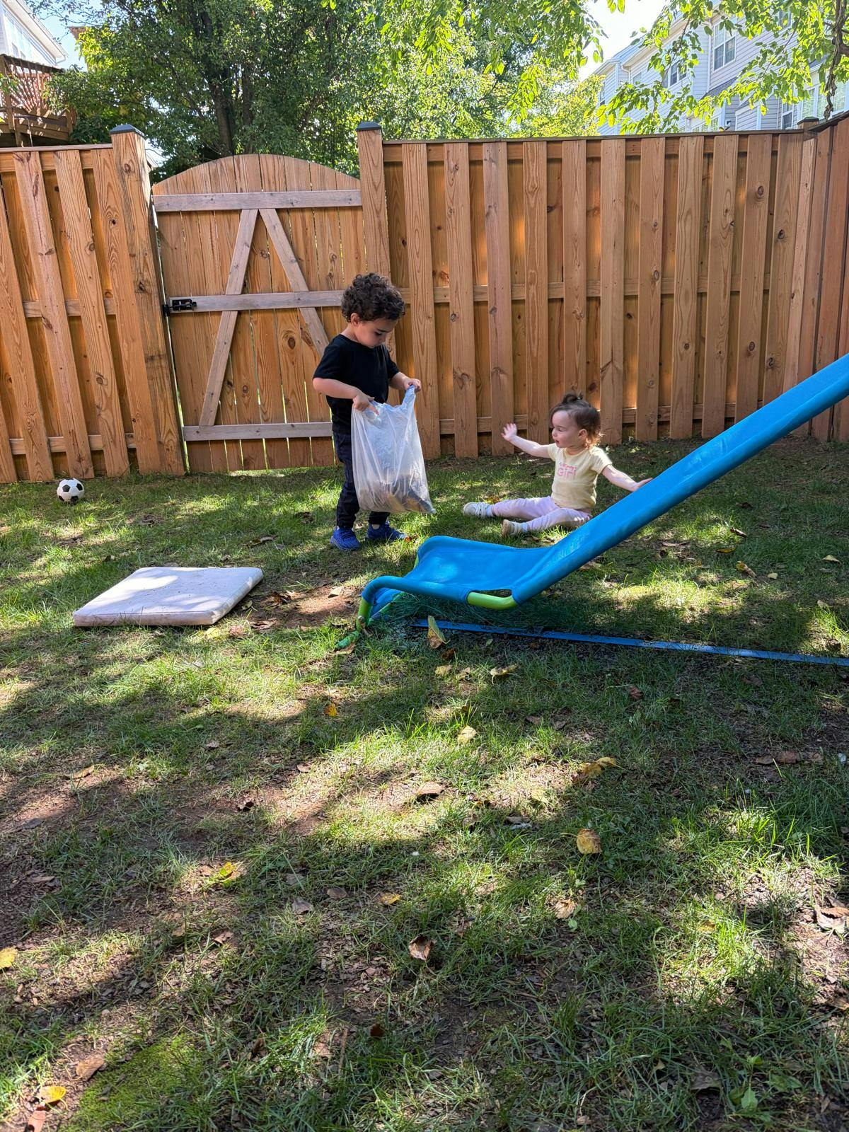 Child holding a bag and a child sitting on the grass by a slide