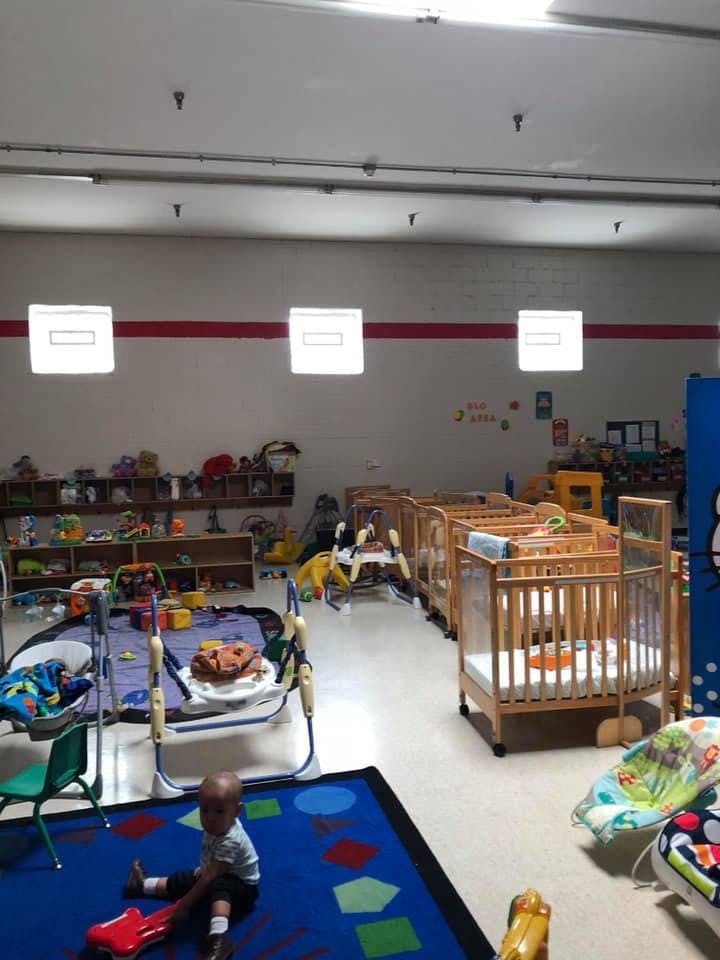 Child sitting on a blue rug in a room with multiple cribs and play equipment