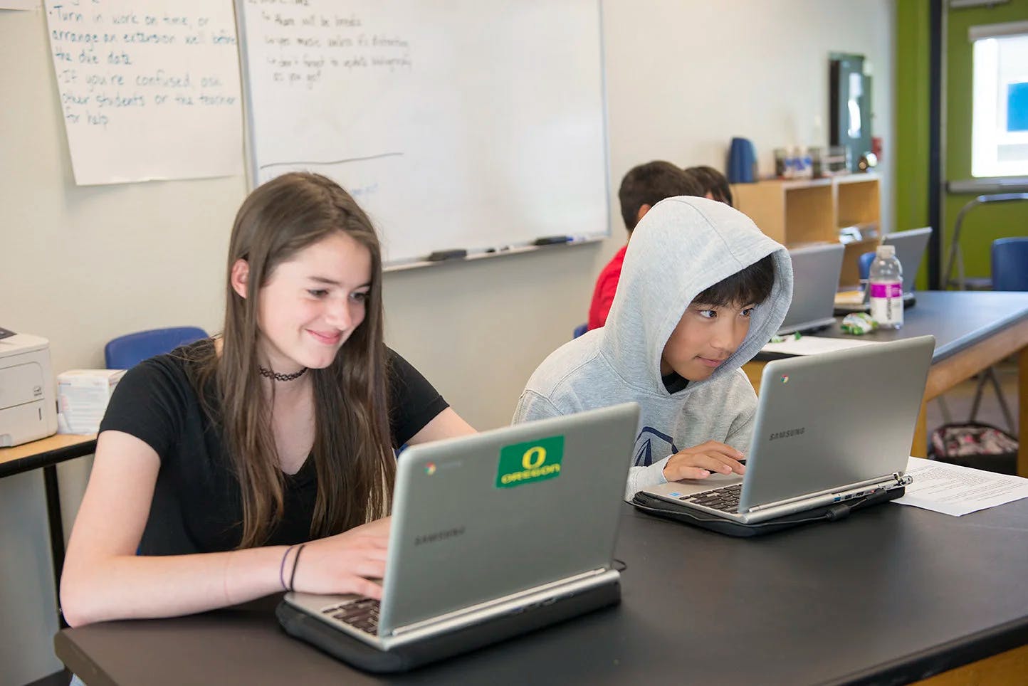 Two teens sitting at a table with laptops in a classroom