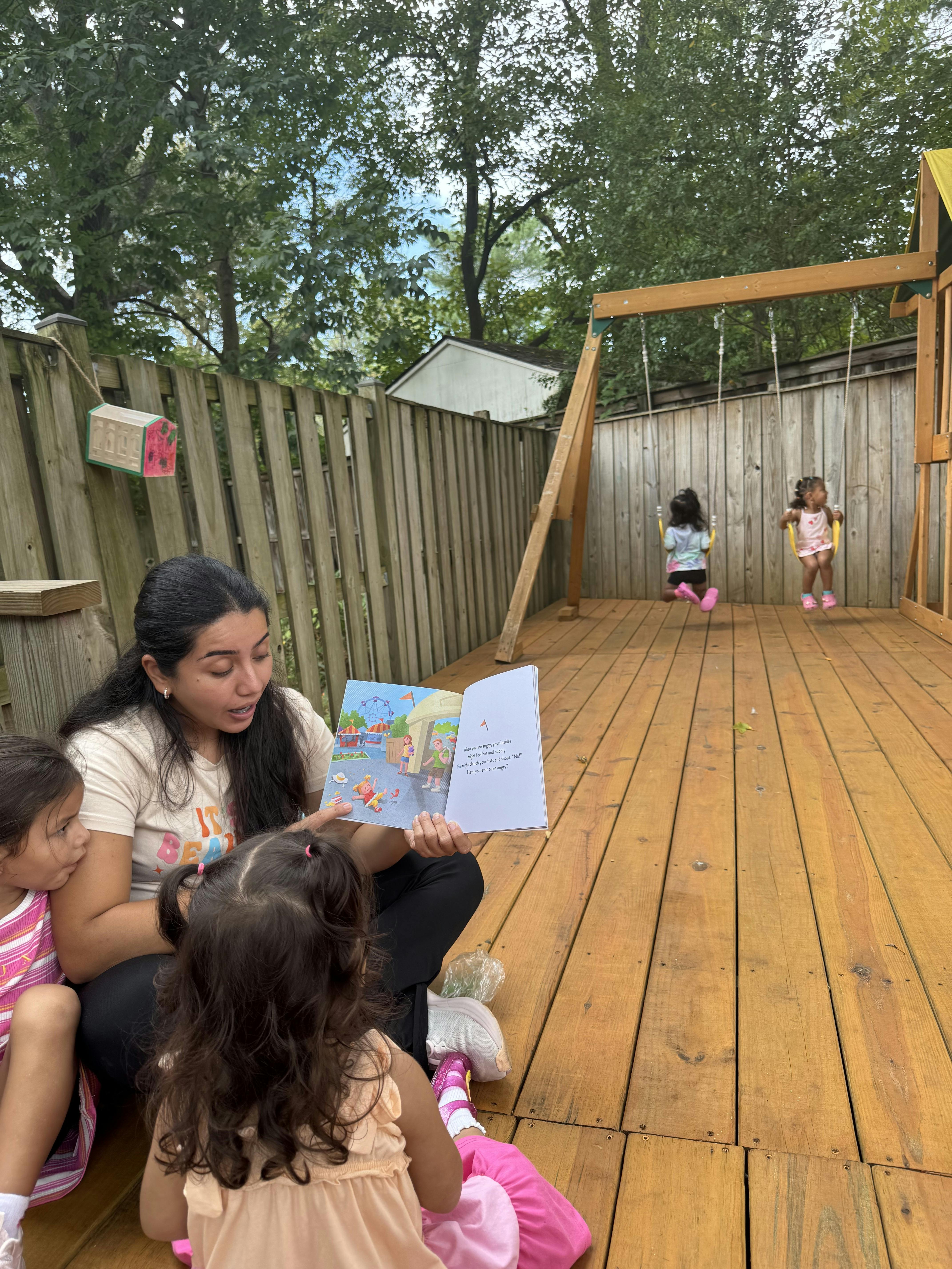 Adult reading a book to two children on a backyard deck with swings in the back
