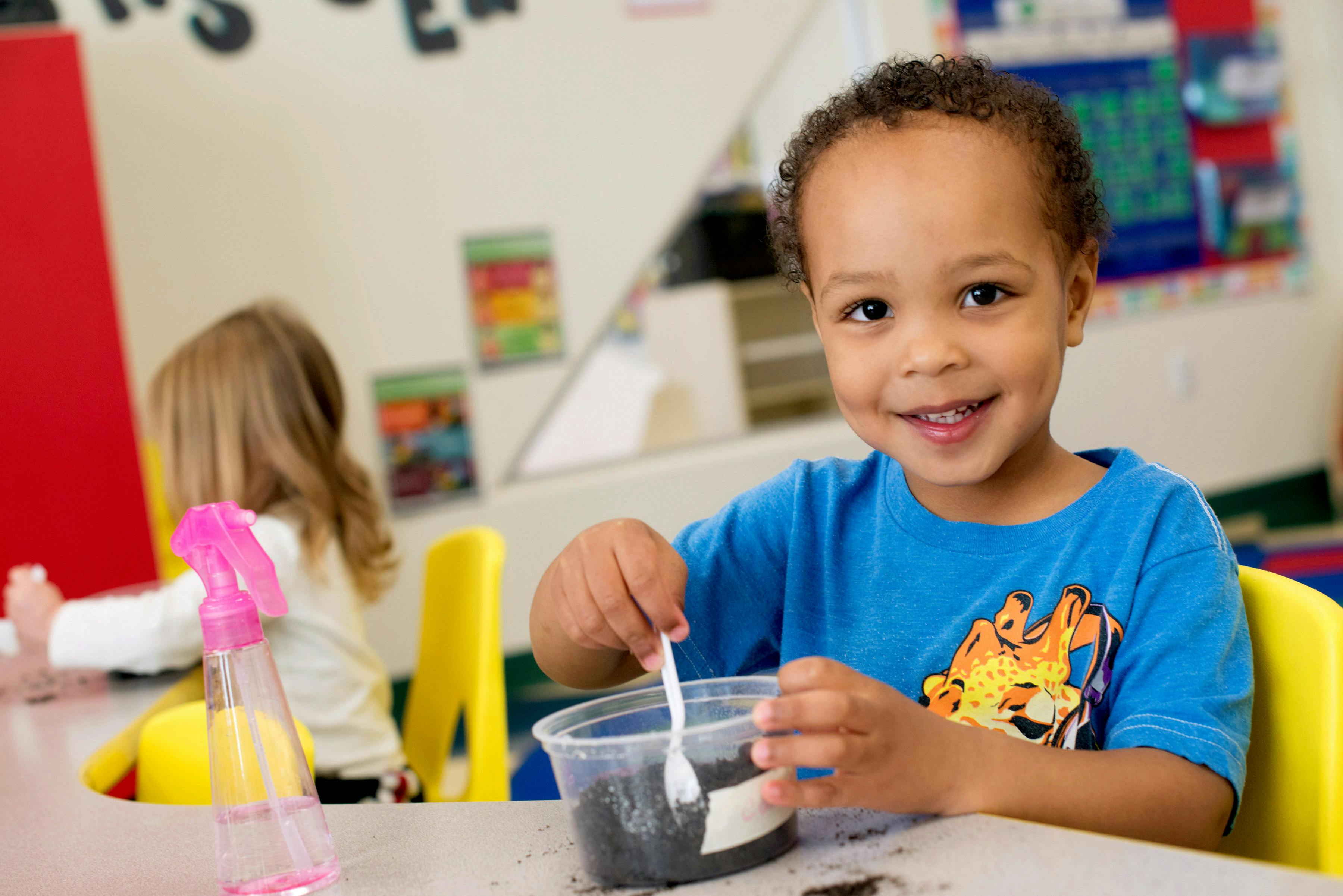 Child stirring dark contents in a clear container with a spoon in a classroom