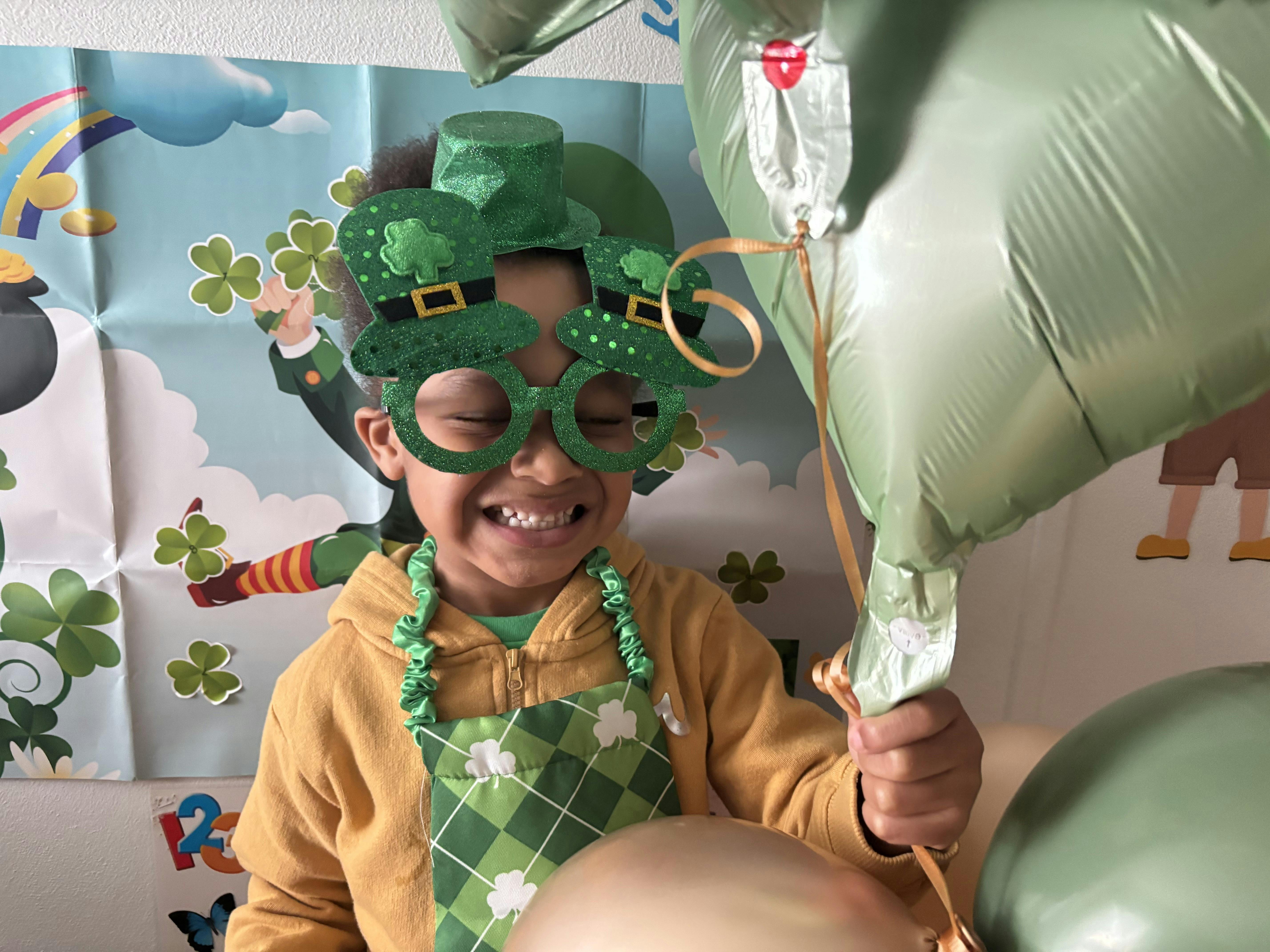 Child in green top hat and novelty glasses holding green balloons