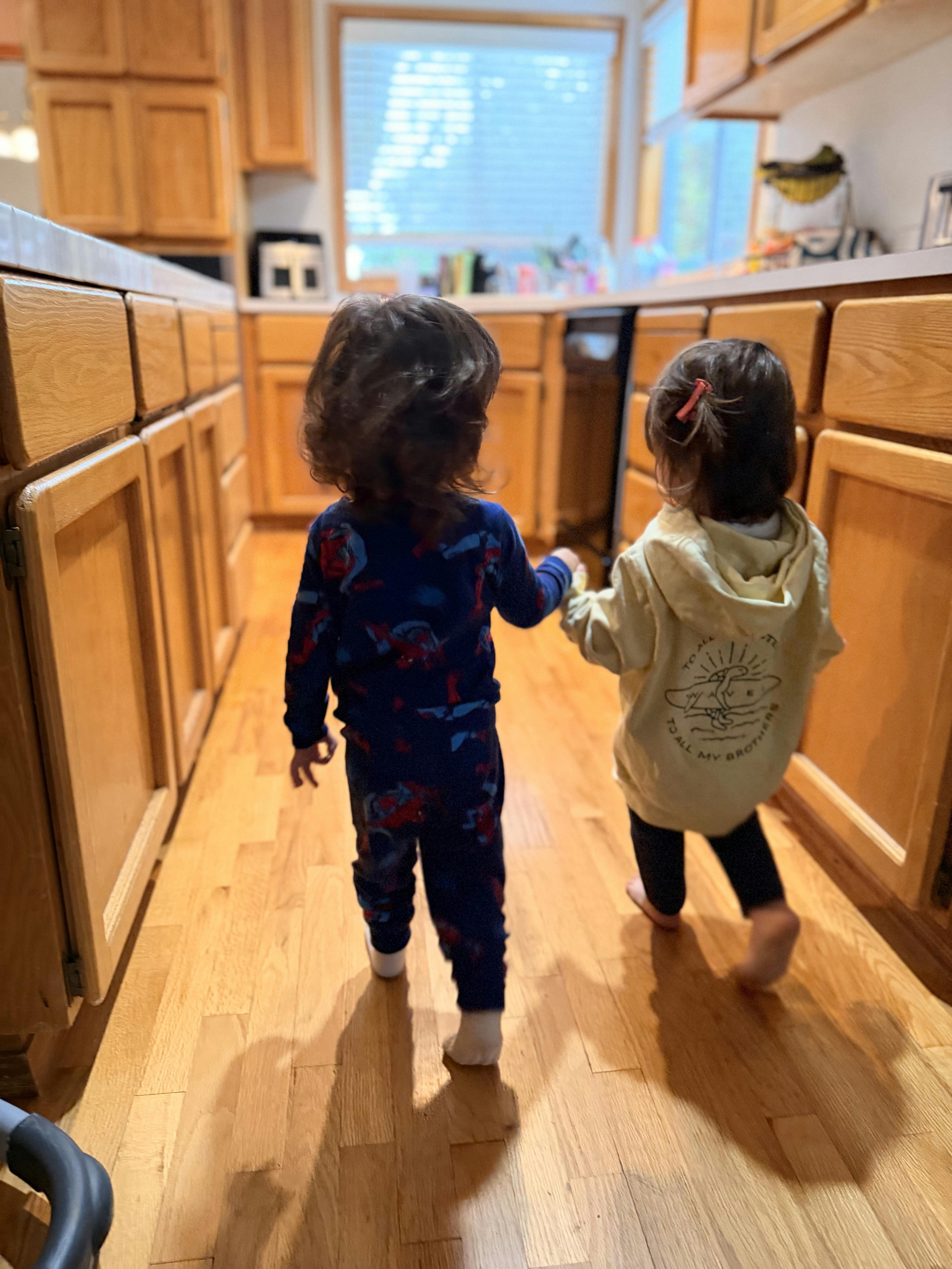 Two children holding hands while walking on a hardwood floor in a kitchen