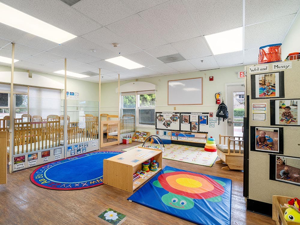 A colorful childcare classroom with cribs, playmats, and developmental toys