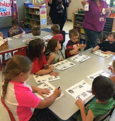 Children sitting at tables with papers and writing utensils in a classroom setting