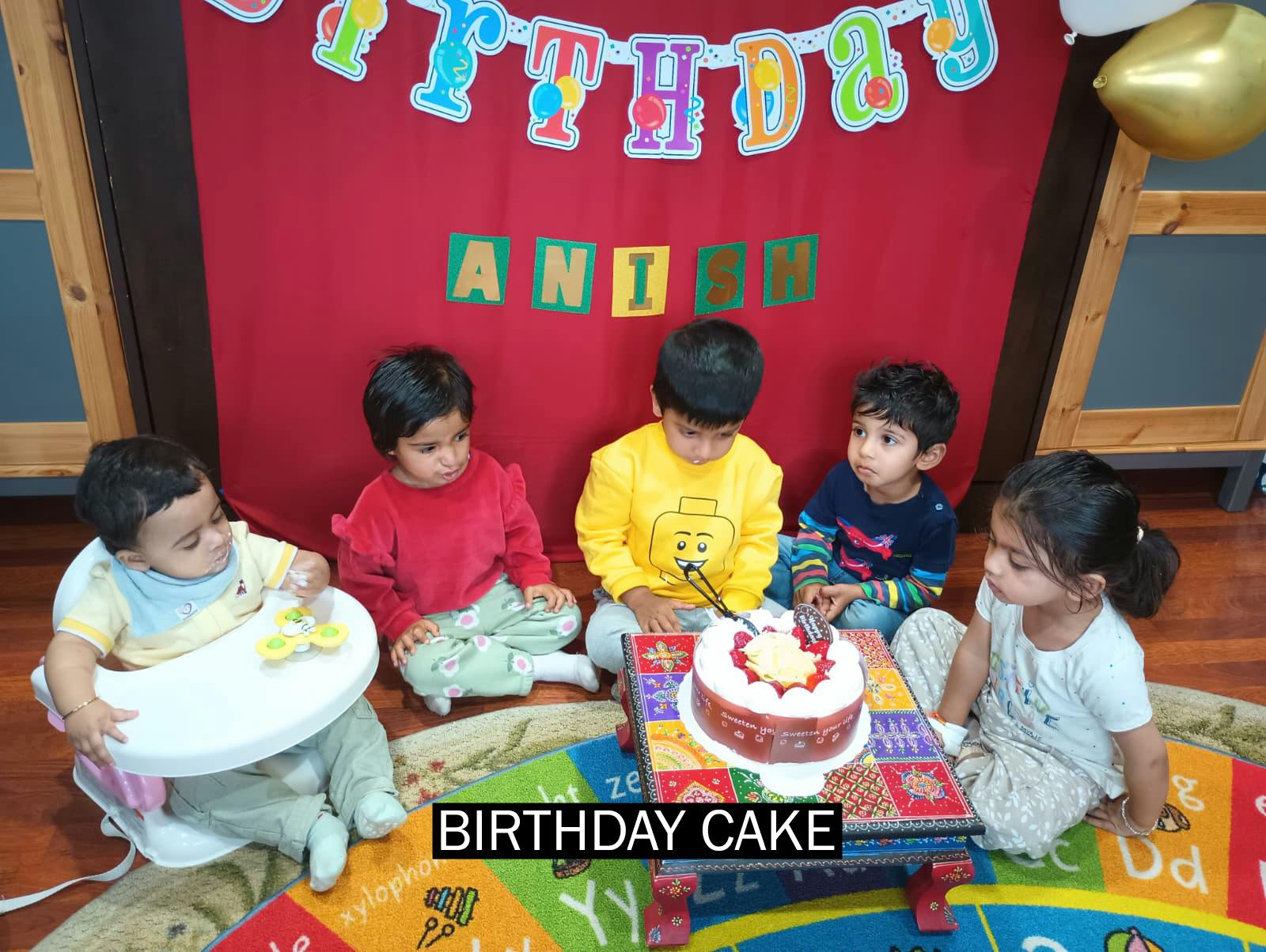 Five children seated around a birthday cake with "BIRTHDAY" banner above