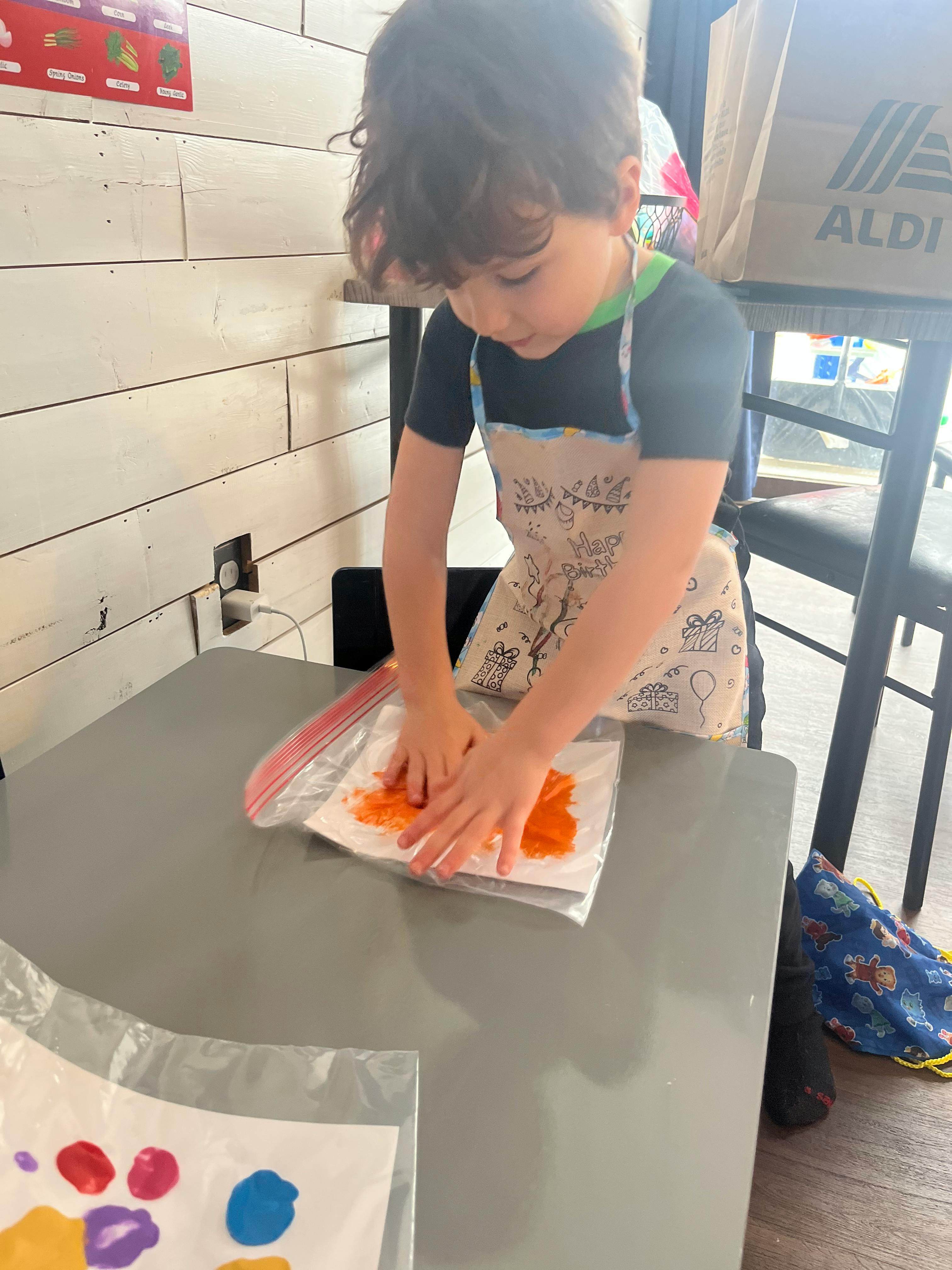 Child wearing an apron pressing hands on paint in a plastic bag