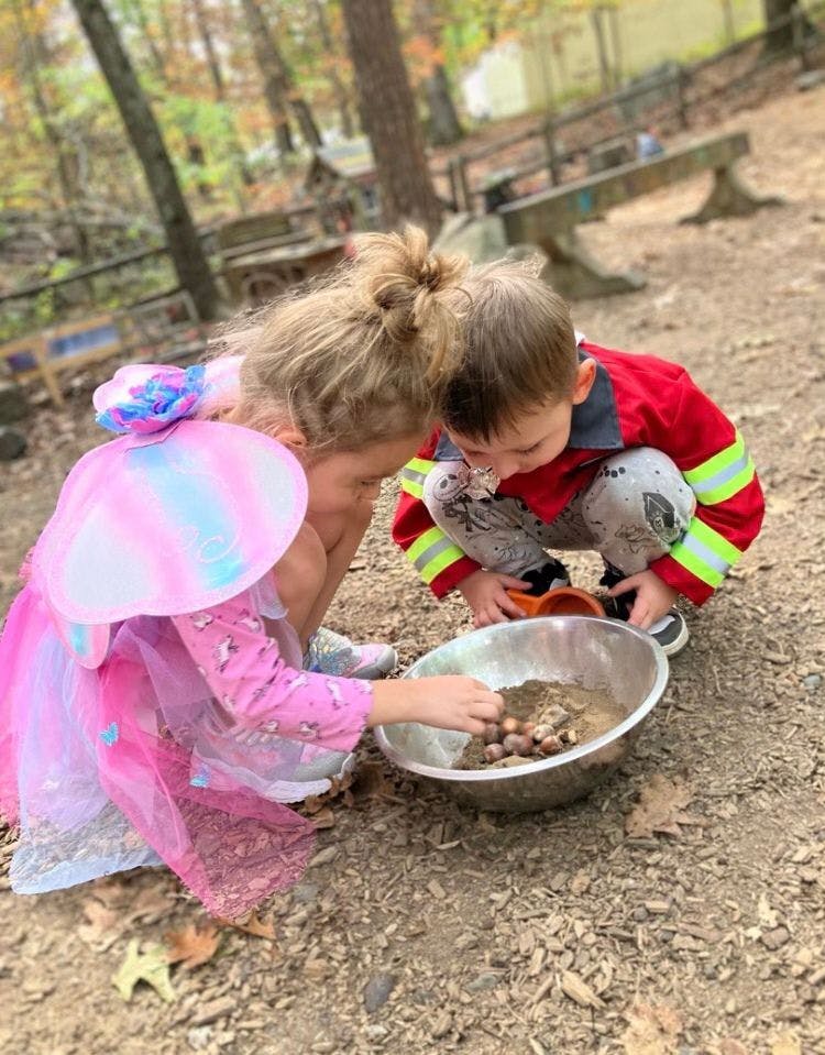 Two children in costumes playing with sand and acorns in a bowl outdoors