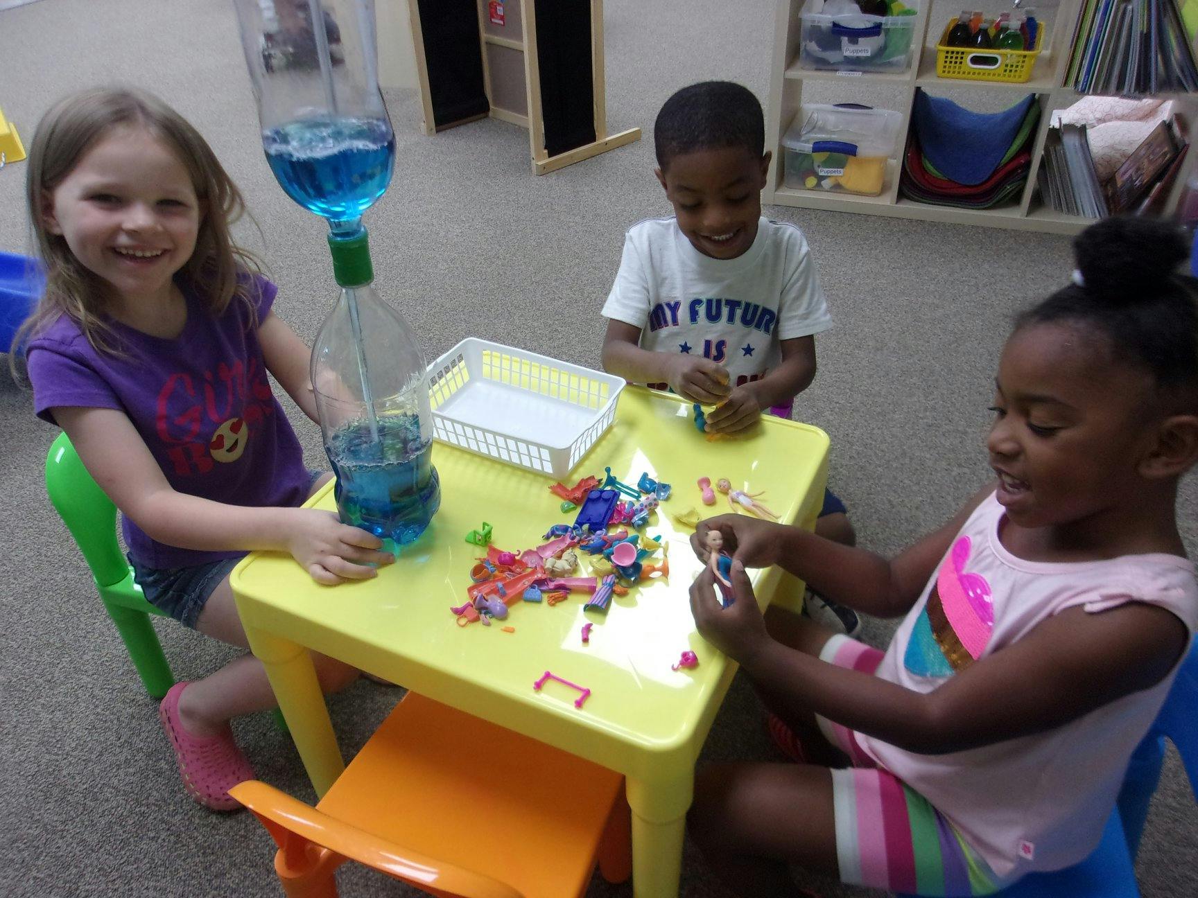 Three children at a table with a water bottle experiment and building toys