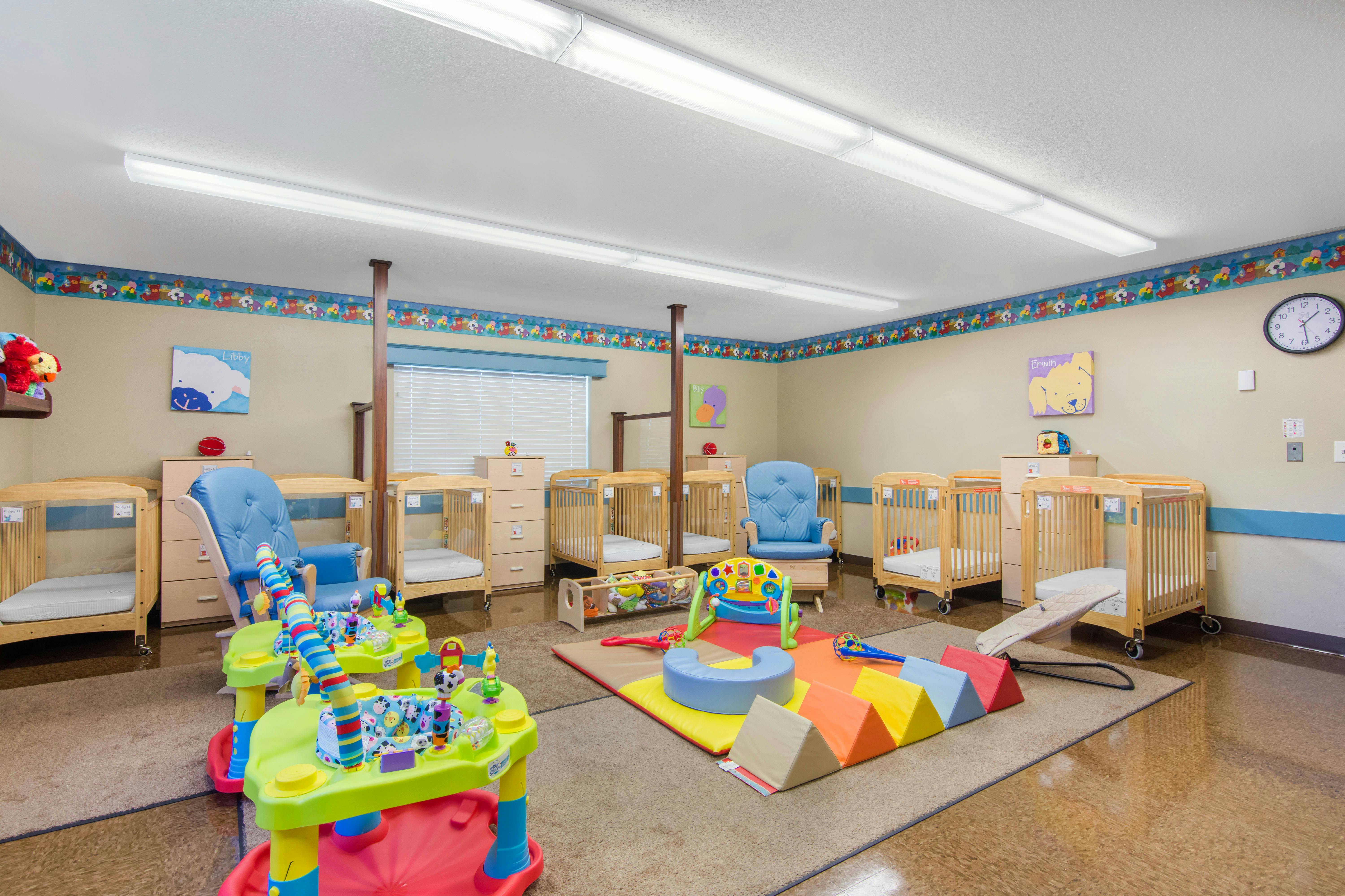 A room with multiple cribs, a blue rocking chair, and colorful play equipment on a rug