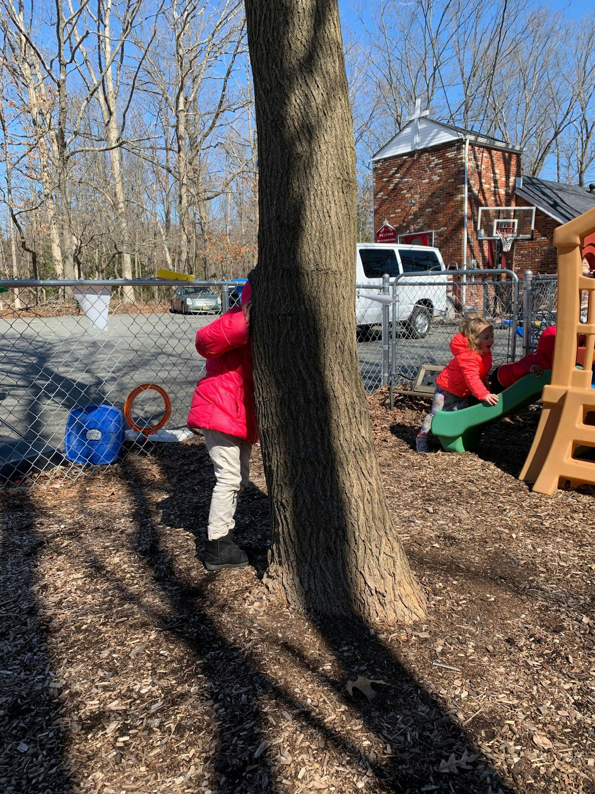 Child in red jacket hiding behind a tree on a playground
