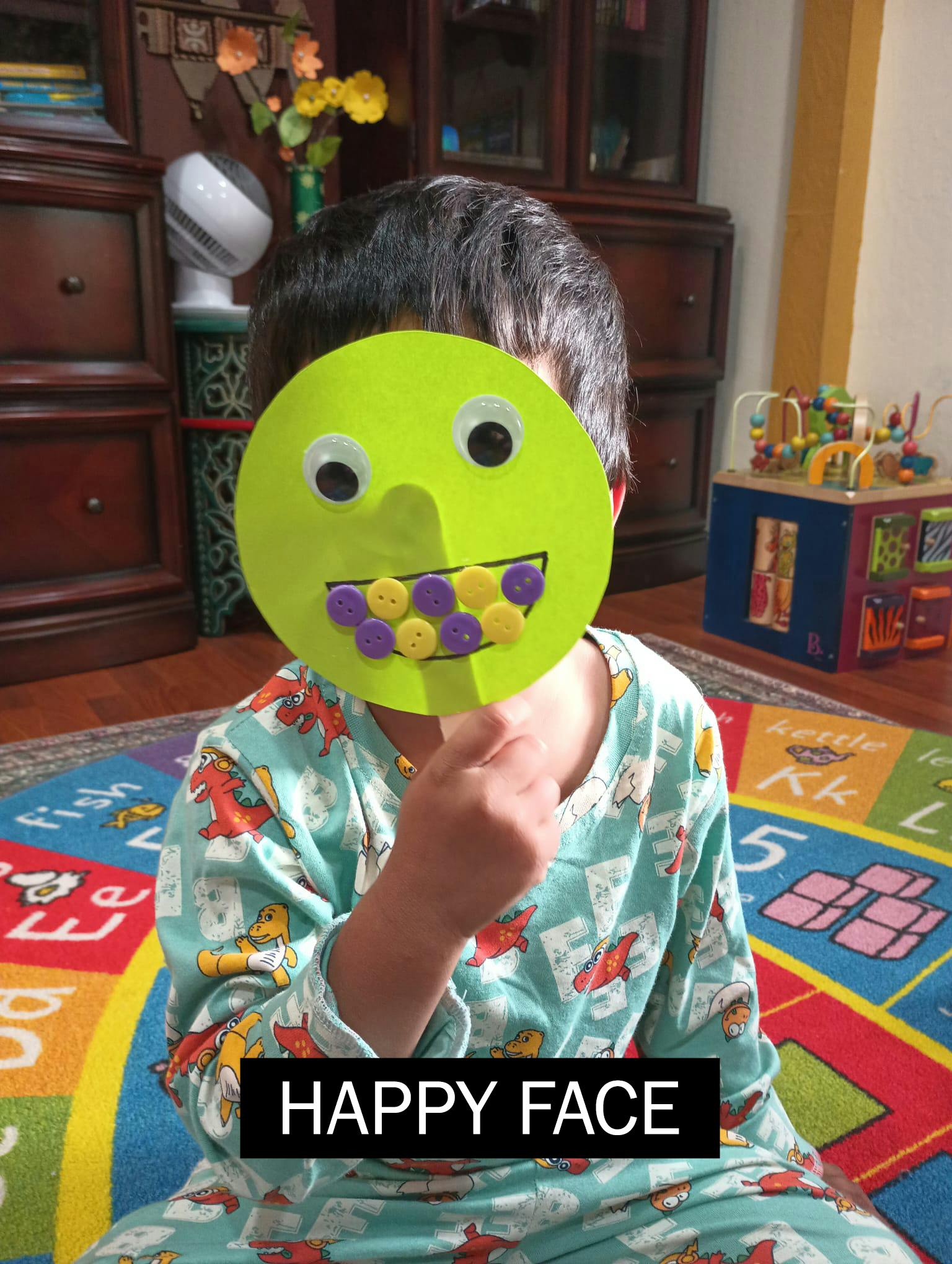 Child wearing dinosaur pajamas, holding a green happy face cutout over their face