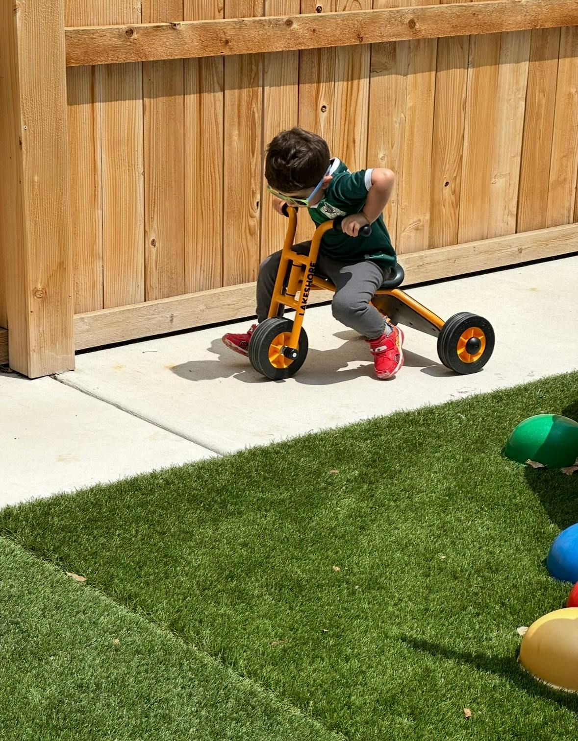 Child in sunglasses riding a tricycle next to a wooden fence