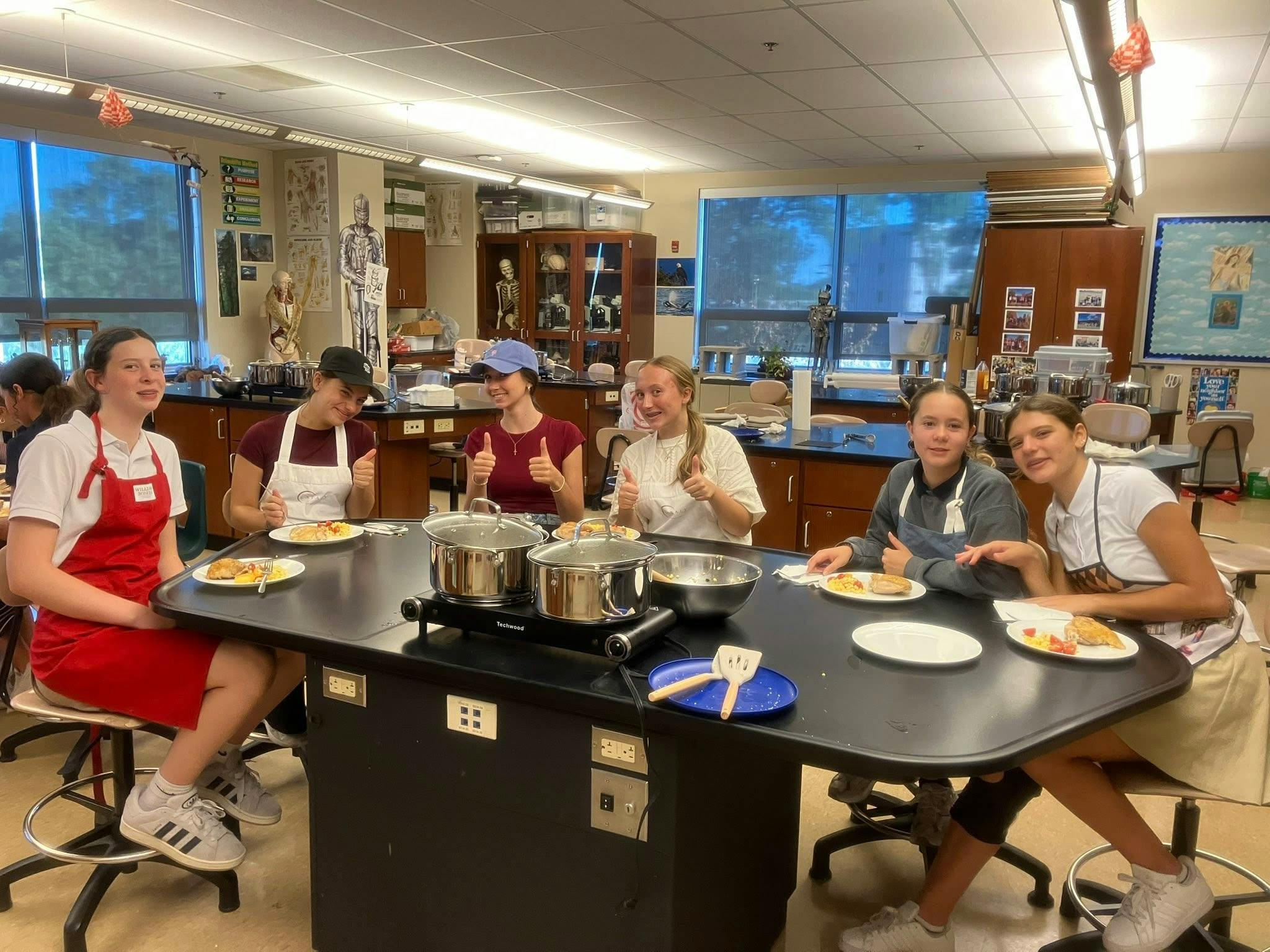 Six teens giving thumbs up around a table with pots and plates of food in a classroom
