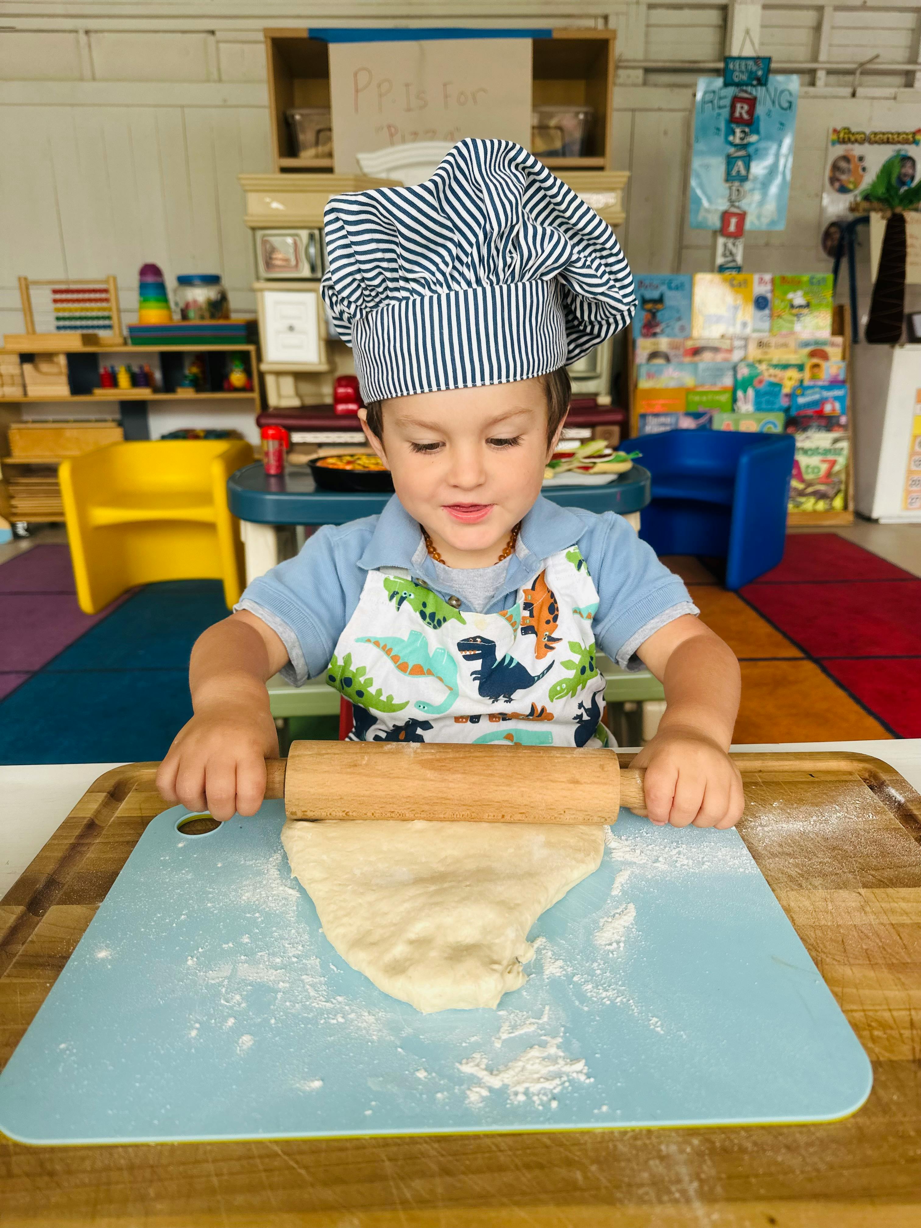 Child in chef hat rolling dough with a rolling pin in a classroom