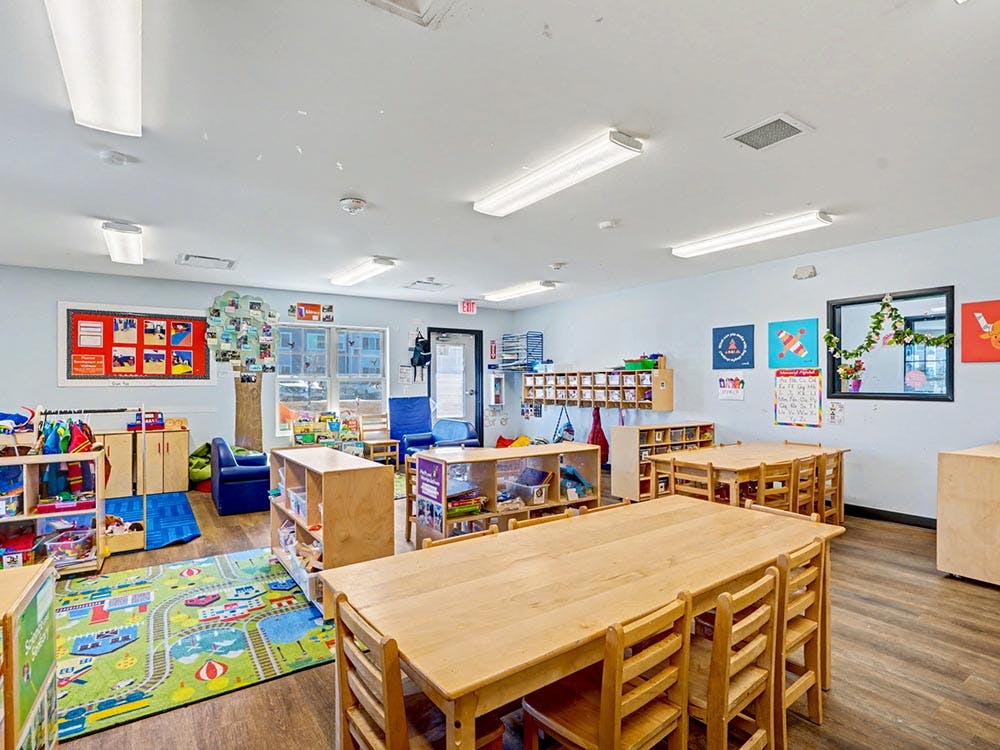 Classroom with small wooden tables and chairs, play rug, and shelves with colorful artwork