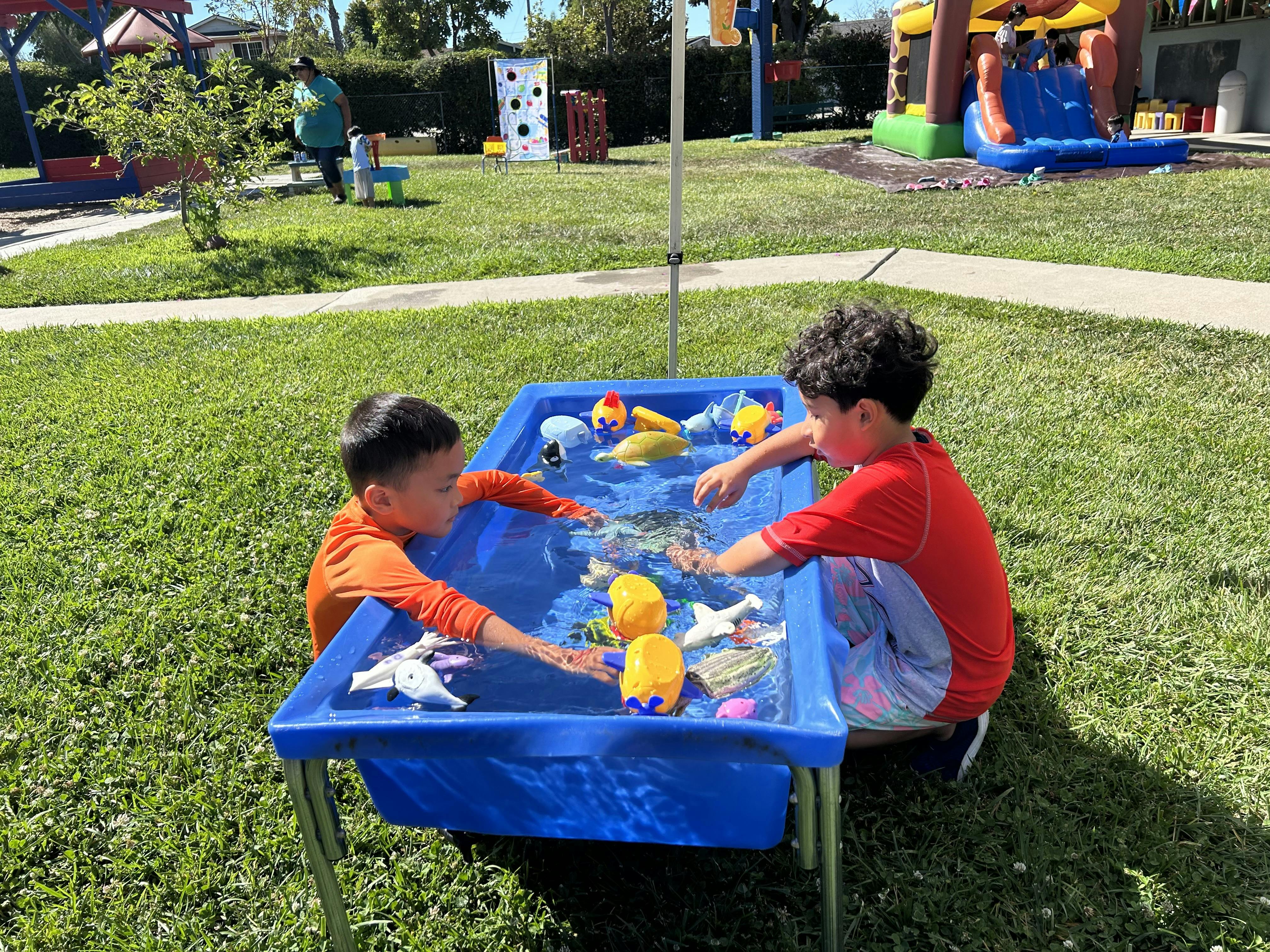 Two children playing with toy figures in a water table on a grassy lawn