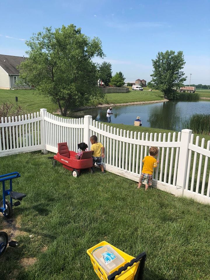 Children playing near a white fence with a pond and person fishing in the background
