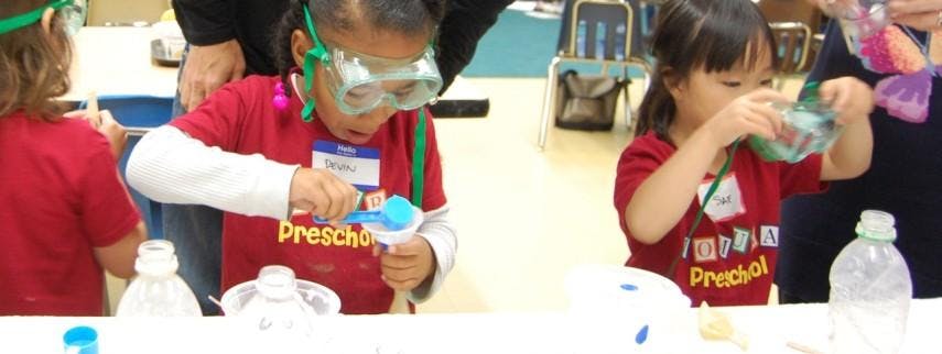 Child in goggles pouring from a scoop, another child pouring from a cup