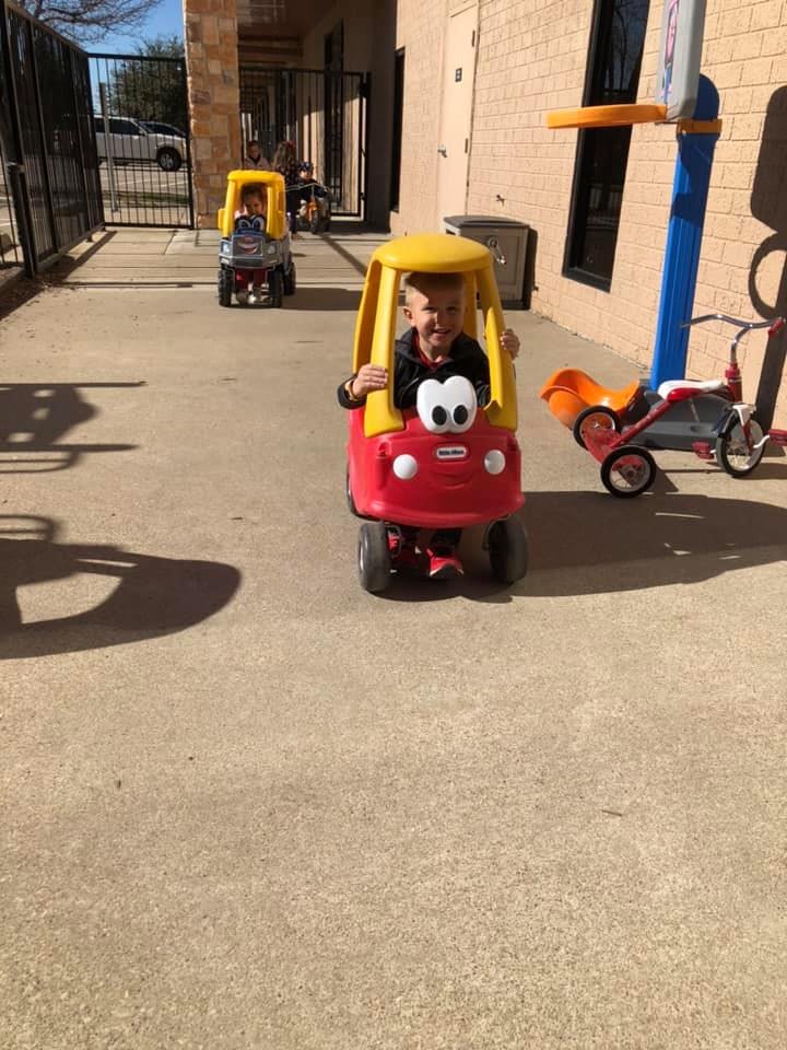 Child in a red and yellow toy car, another child in a toy car behind