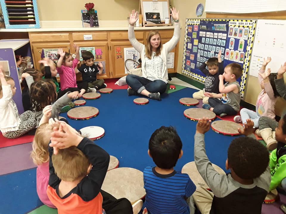 Adult and children sitting in a circle with hands raised in a classroom