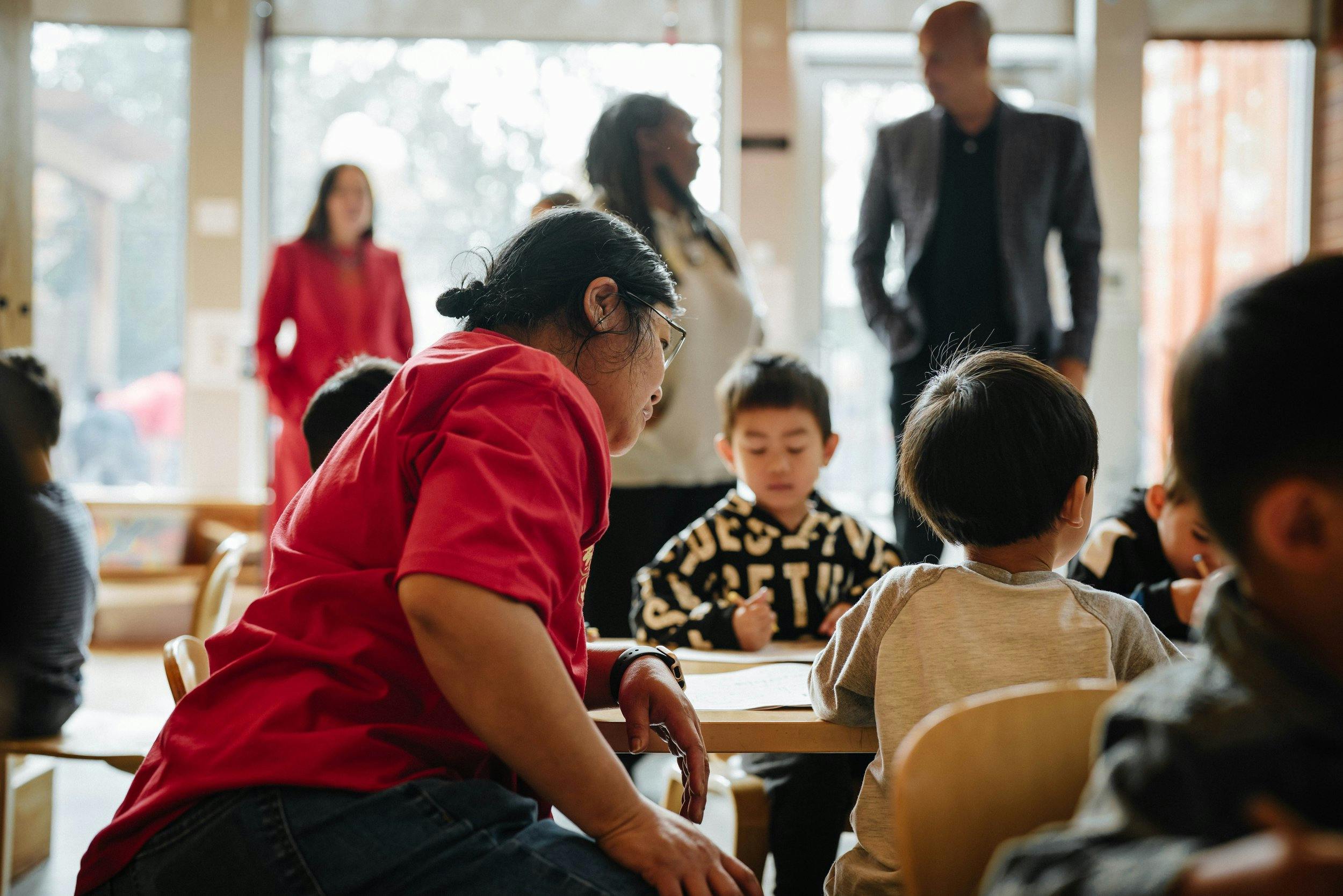 Adult in red shirt looking at children sitting at a table in a classroom