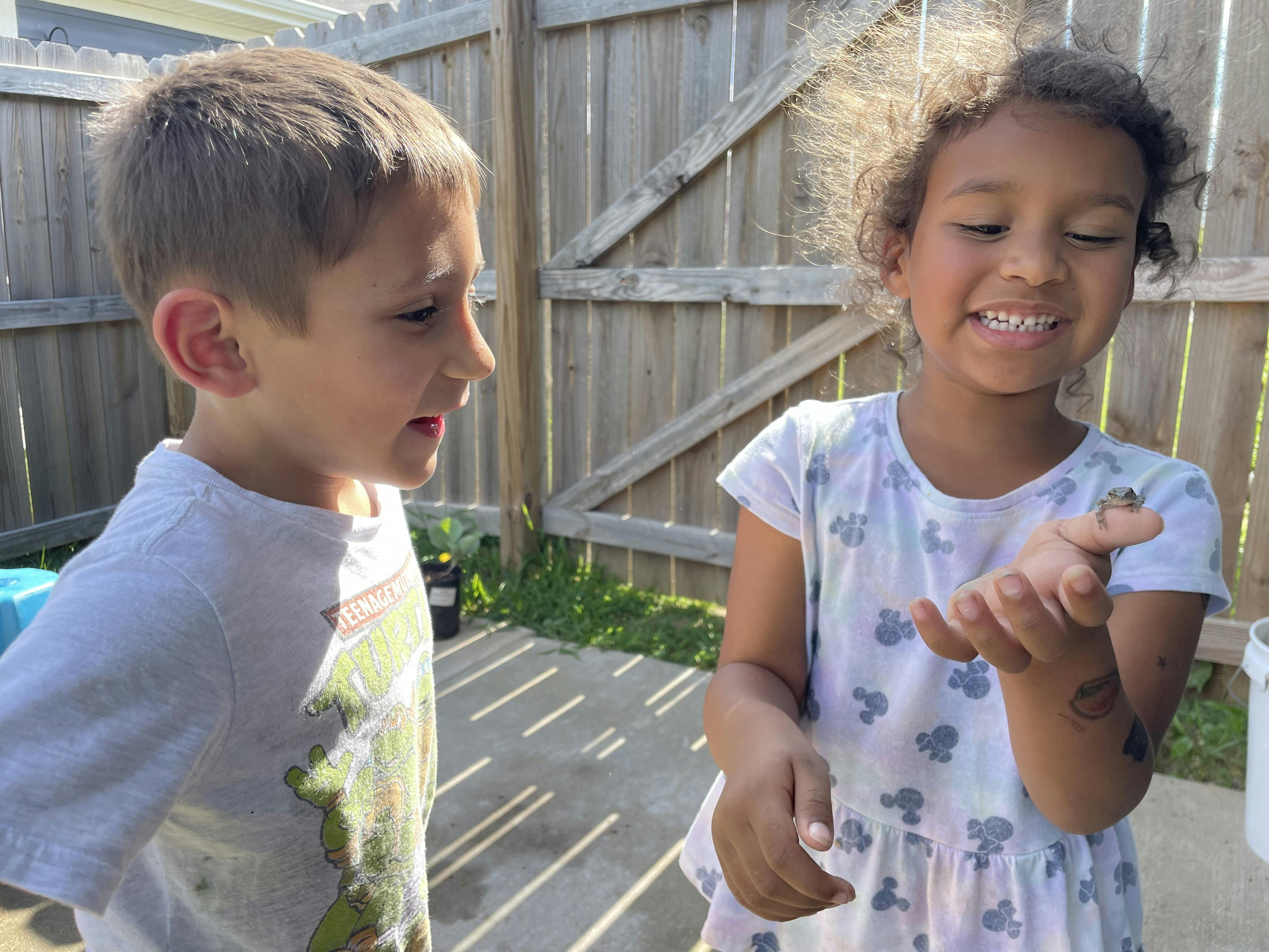Child holding a small frog on her finger, another child watches