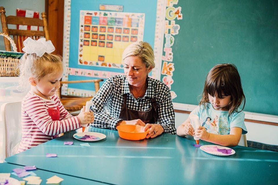 Person and two children doing crafts at a table in a classroom