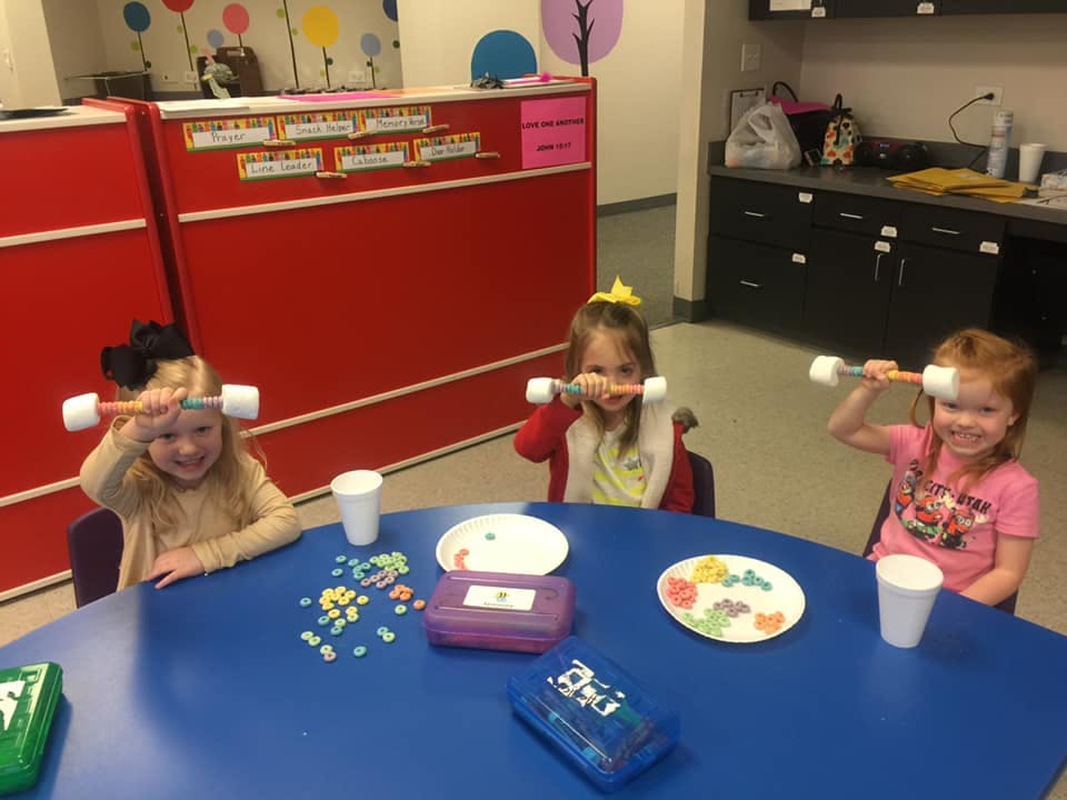 Three children sitting at a table holding toy dumbbells