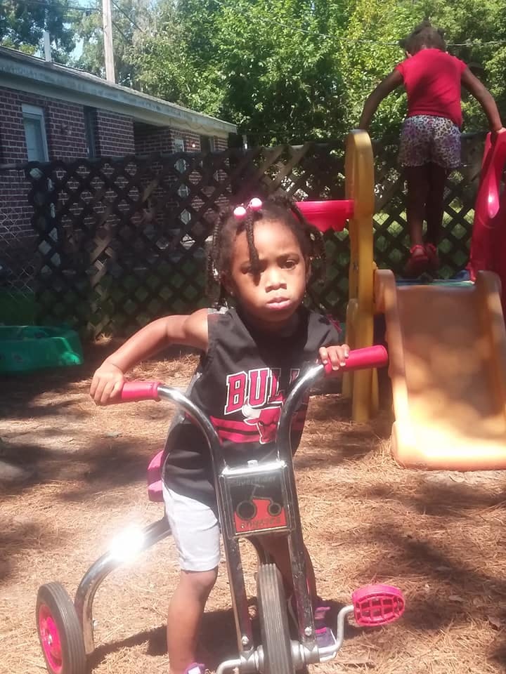 Child on a tricycle with another child on a playground slide in the background