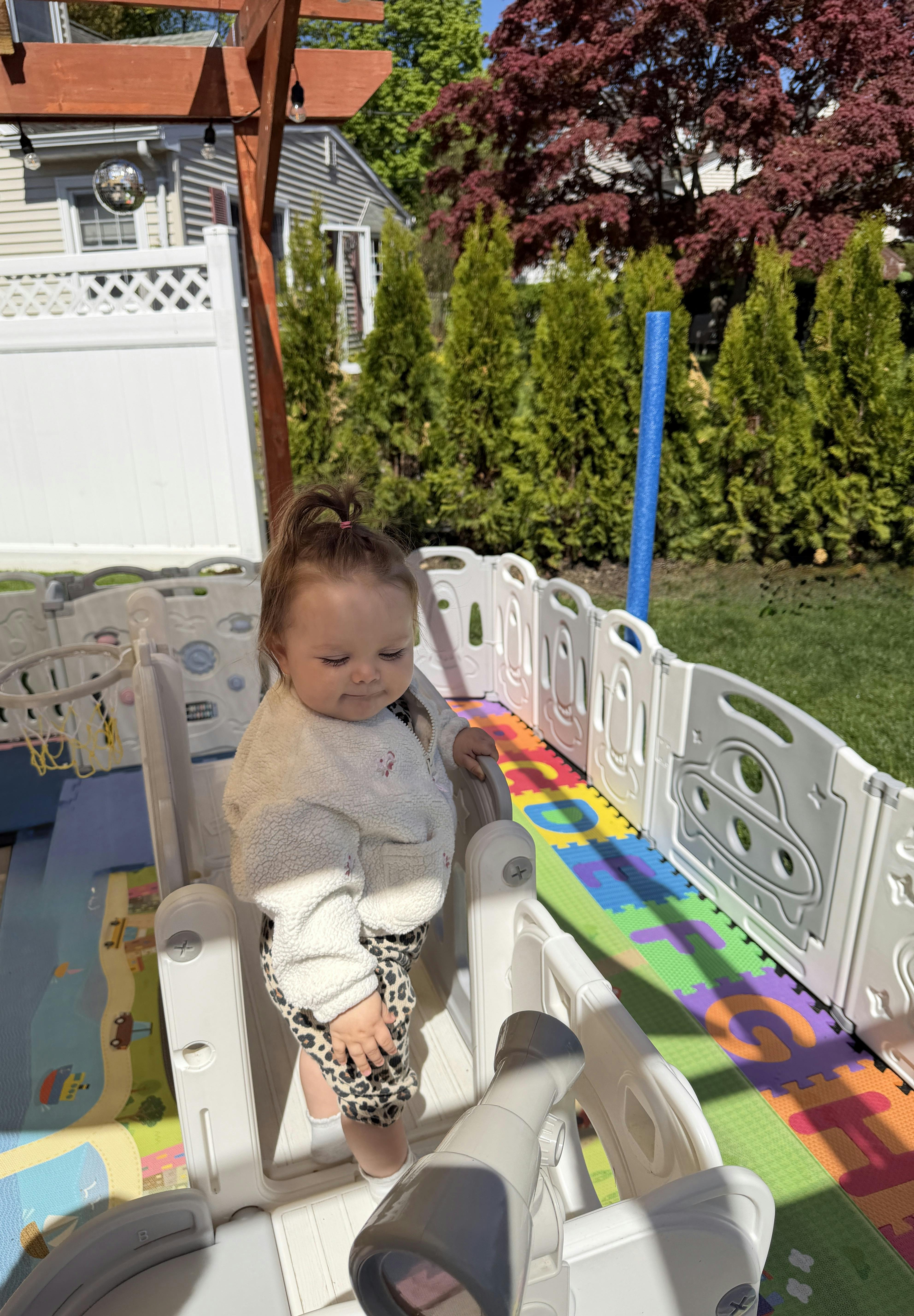 Child standing in a playpen on a colorful mat outdoors