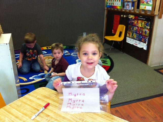 Child at a table with a paper that says "Myra" with other children in the background