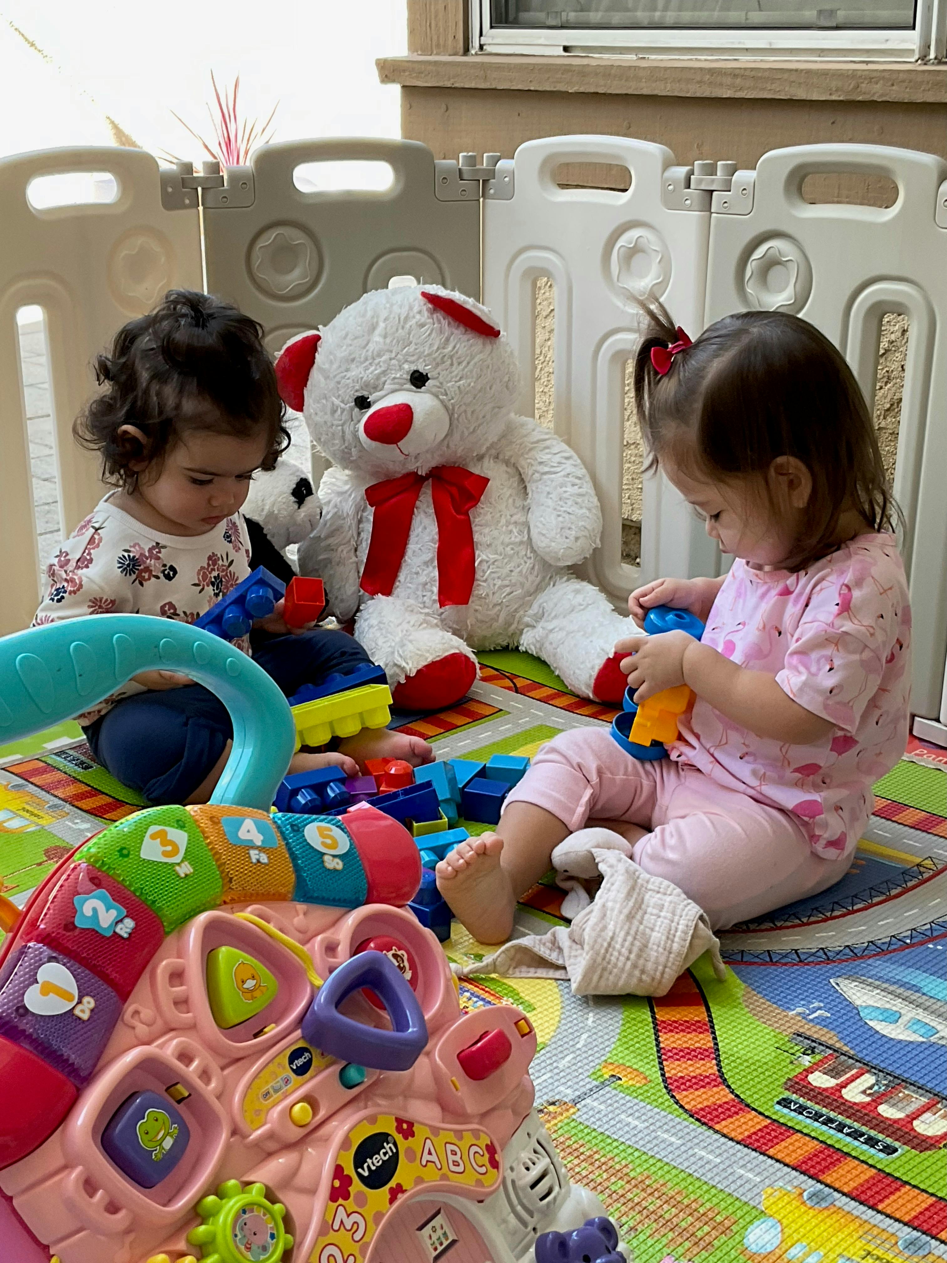 Two children playing with toys and a teddy bear on a colorful mat