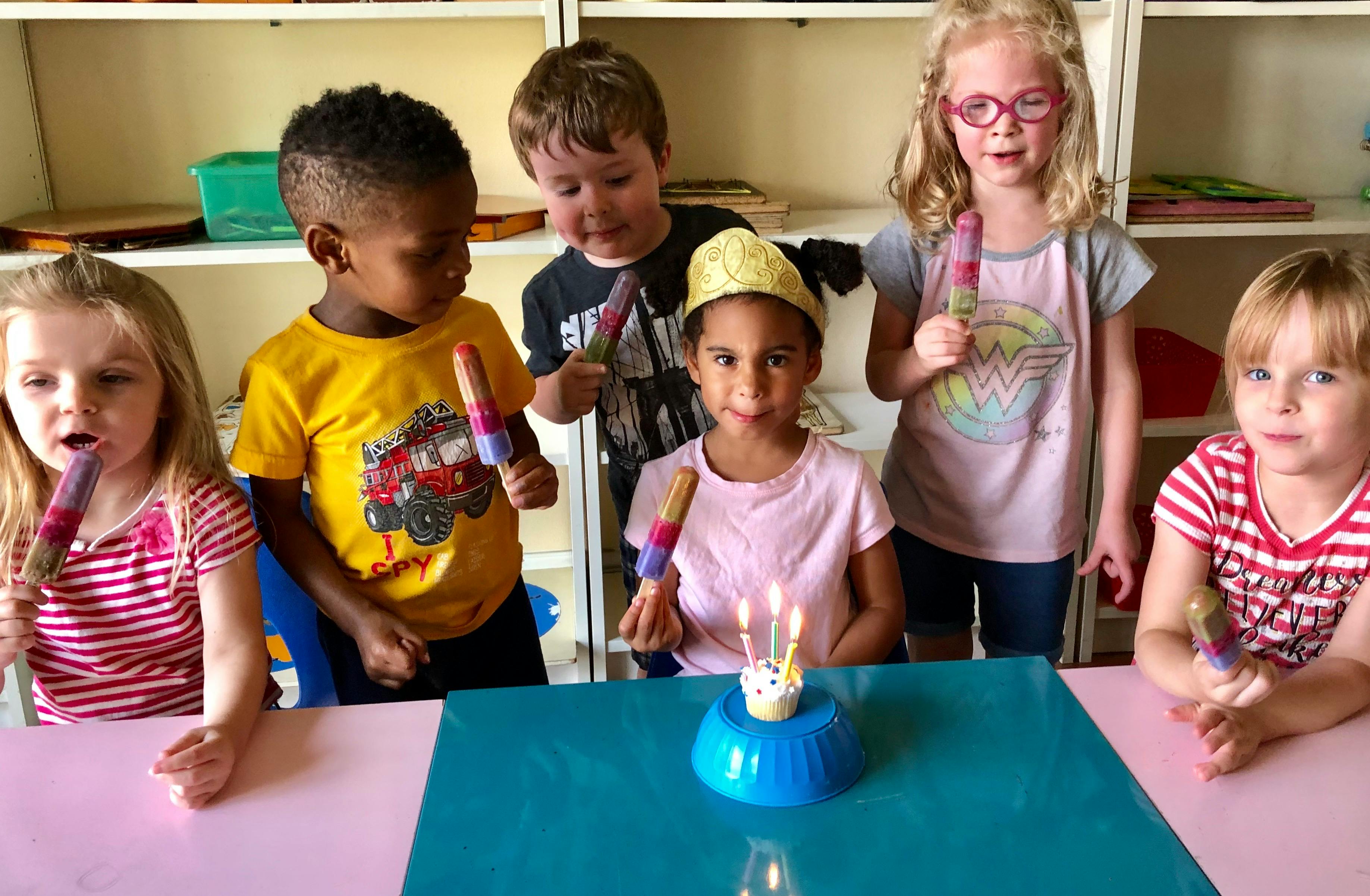 Children holding popsicles around a small cupcake with candles