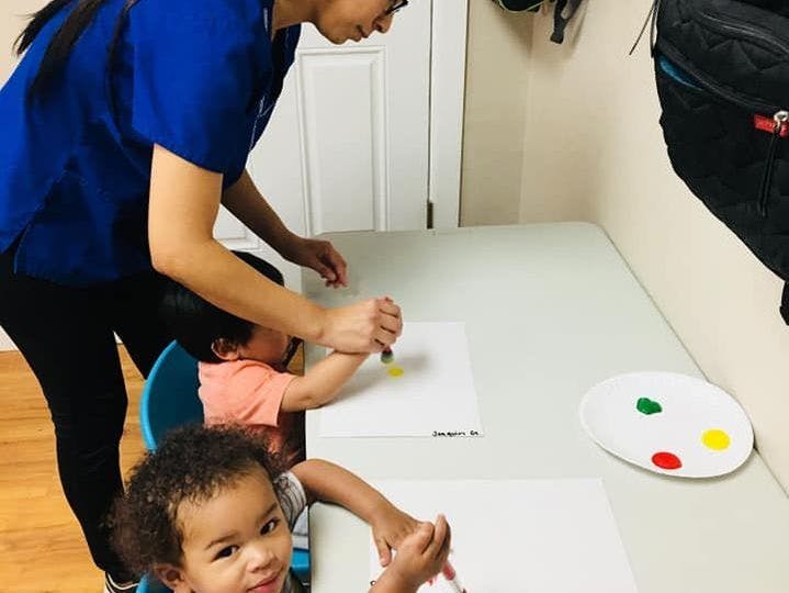 Person helping a child paint at a table with another child nearby