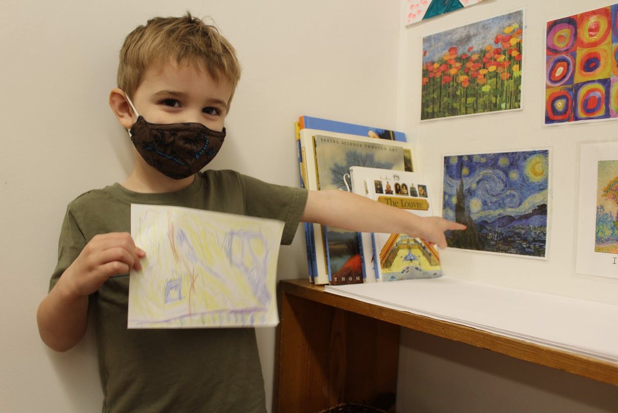 Child in a mask holding a drawing and pointing to artwork on a wall